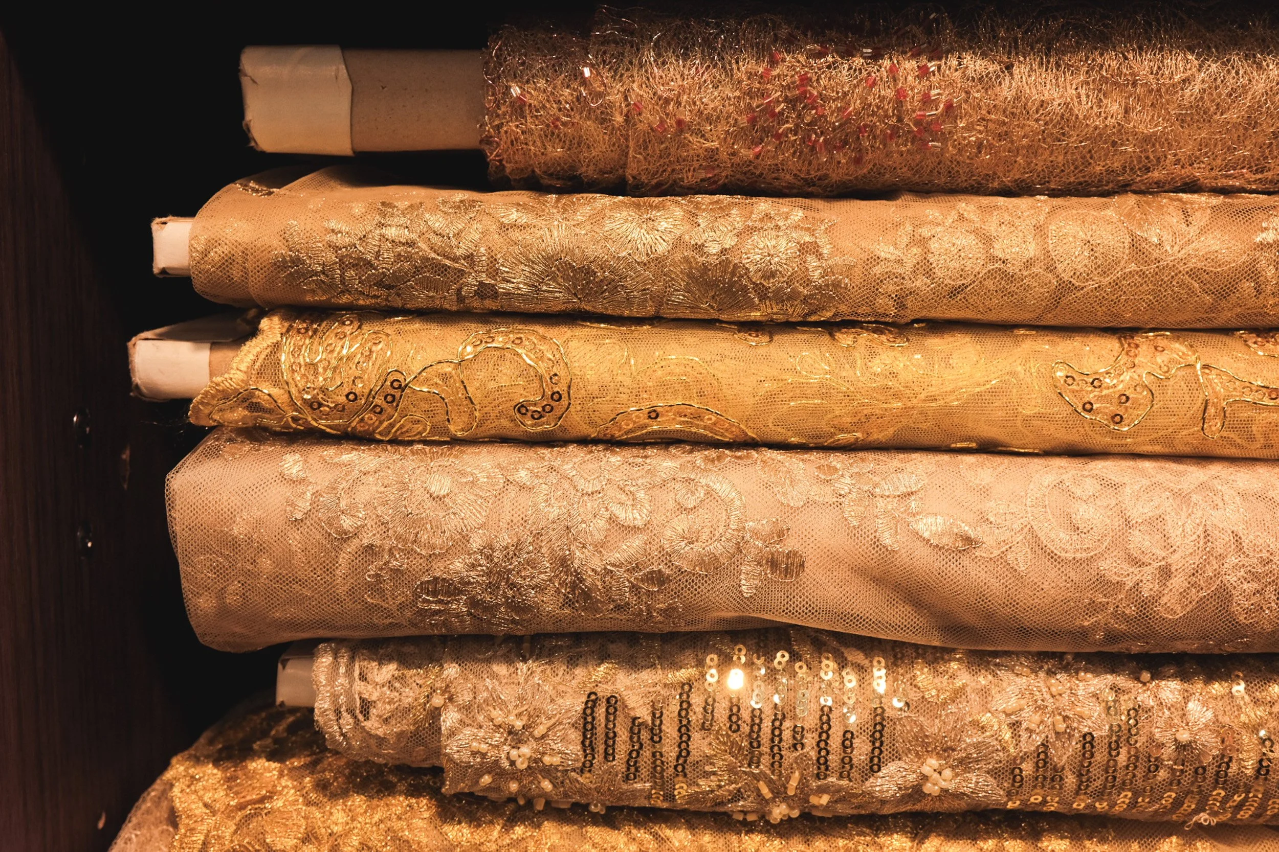 Stacked spools of gold and beige fabric with intricate patterns, sequins, and embroidery.