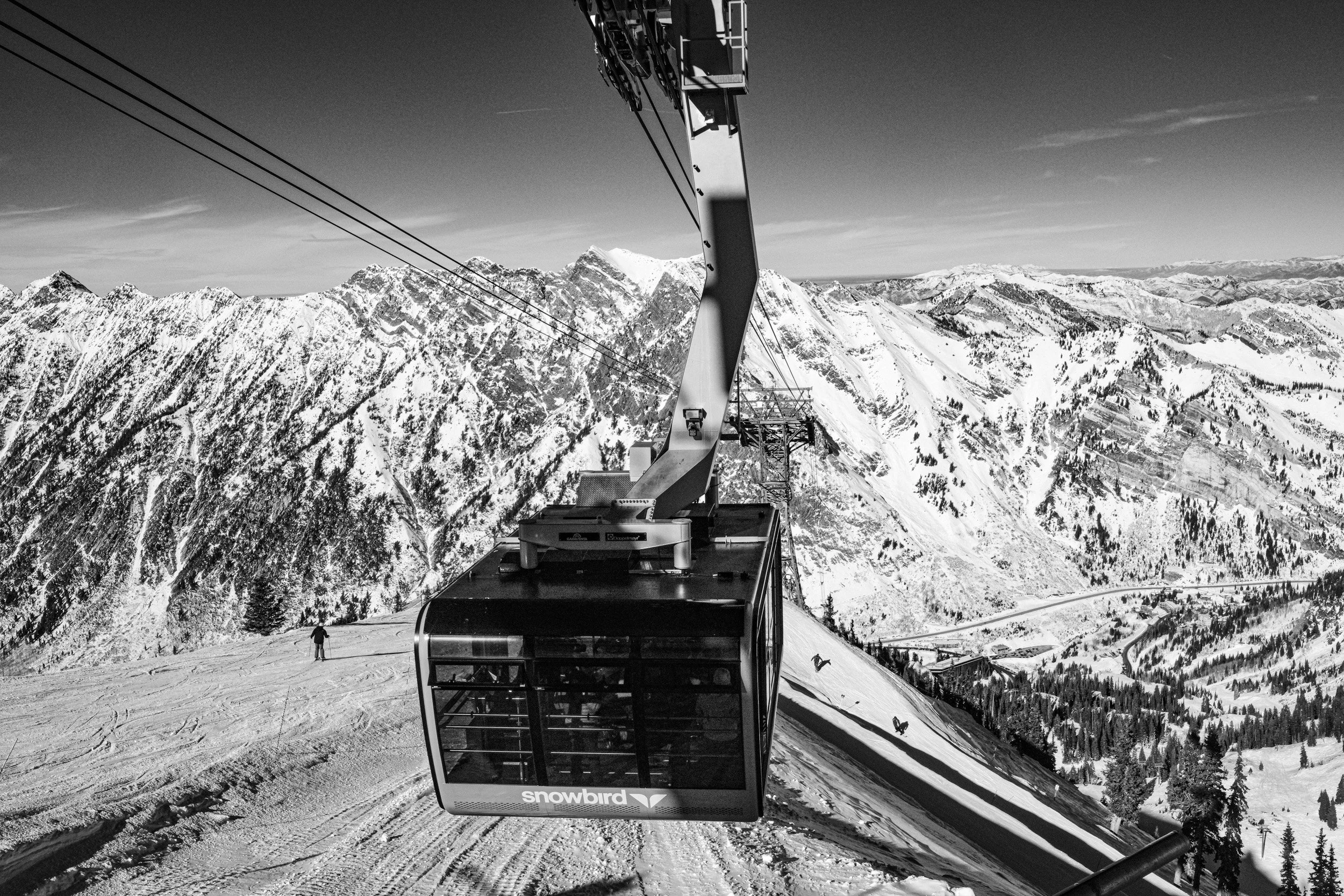 A snow-covered mountain landscape with a ski lift or gondola in the foreground and skiers on the slope. The scene is captured in black and white.