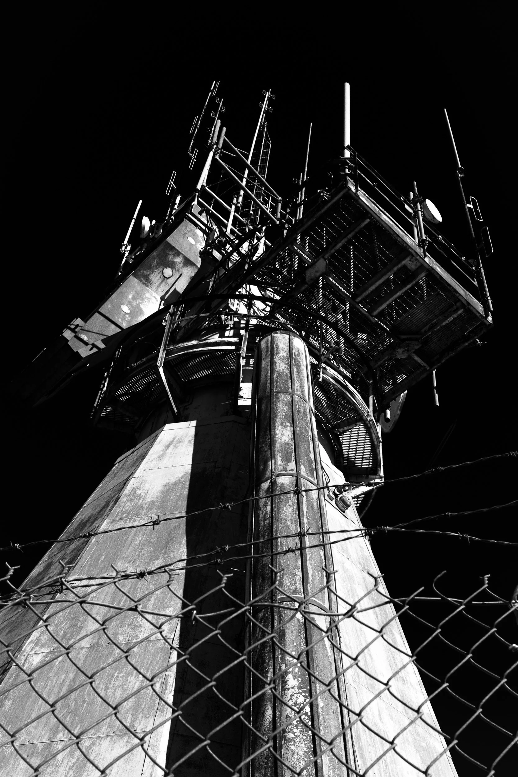 A black and white photo taken from the ground looking up at a communications tower behind a chain-link fence.