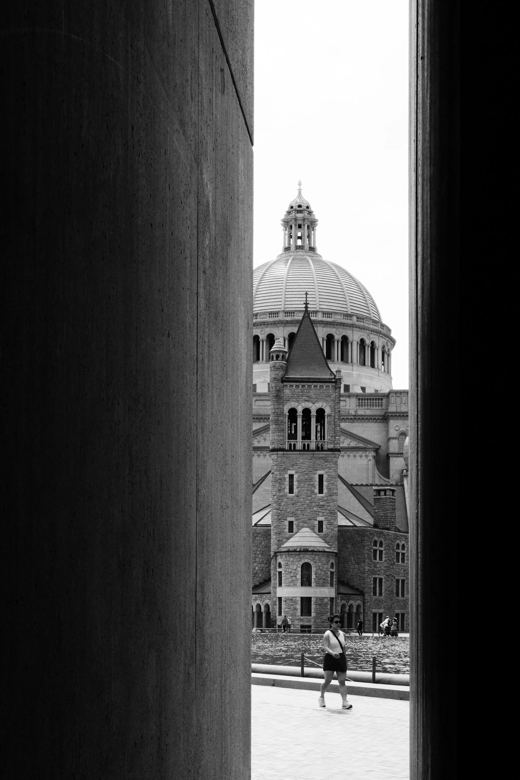 View of a historic building with a large dome seen through a narrow opening, with people walking near a river in the foreground.