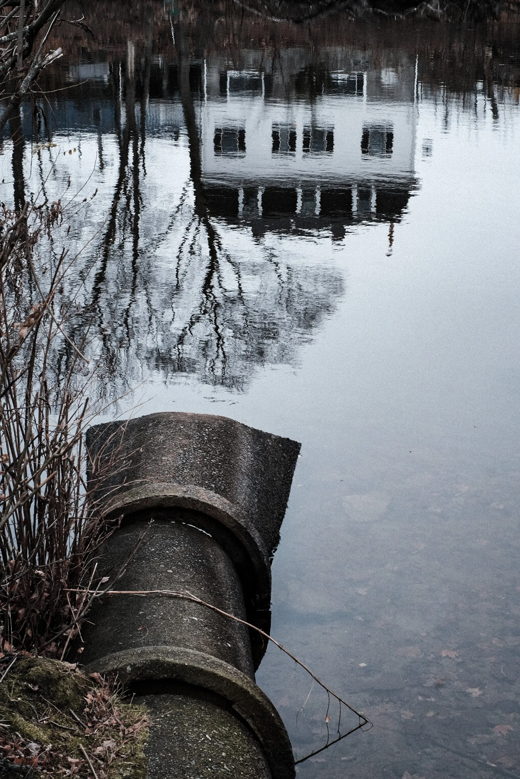 A line of large concrete pipes lying along the bank of a river or lake with reflections of trees and a white building with multiple windows and a chimney in the water