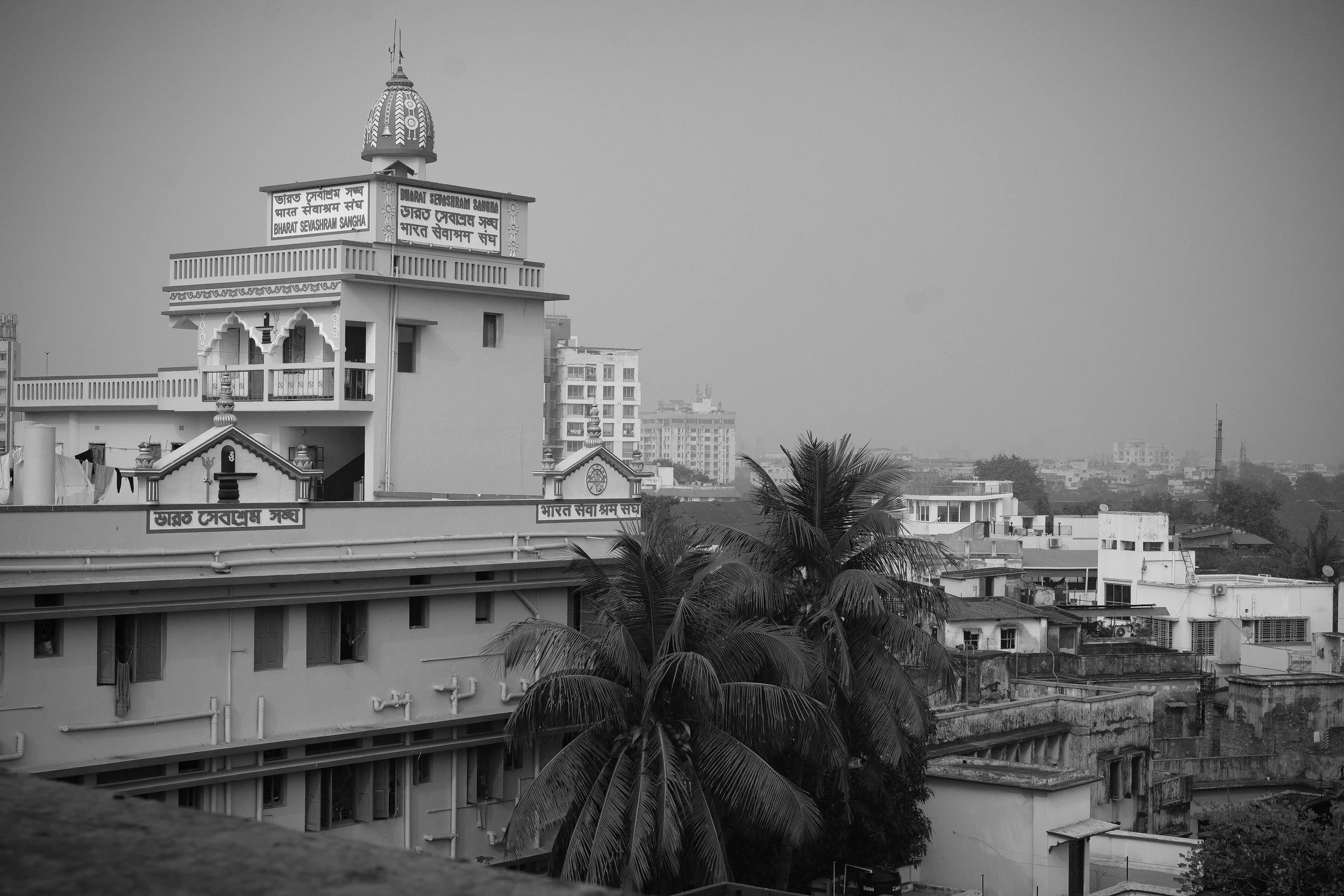 Black and white photo of a cityscape with a building featuring Hindu architectural details, signs in Hindi and English, and multiple buildings in the background.