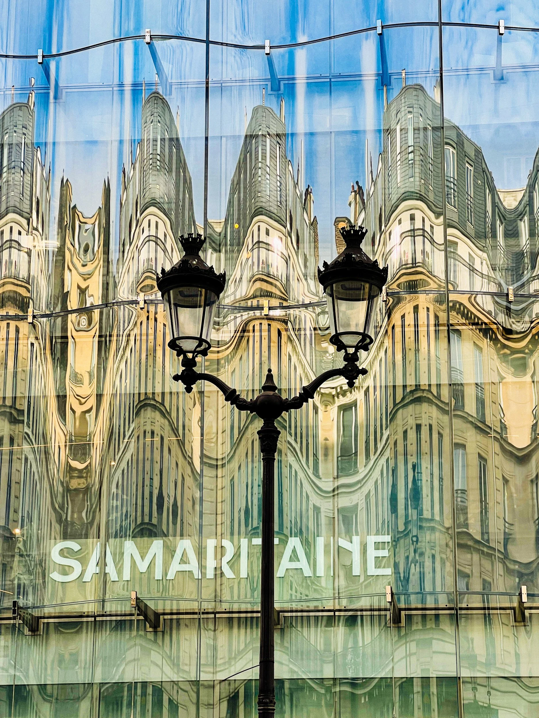 Modern building with reflective glass facade distorting the reflection of older style buildings and a streetlamp in front, with the sign 'SAMARITAINE' visible.