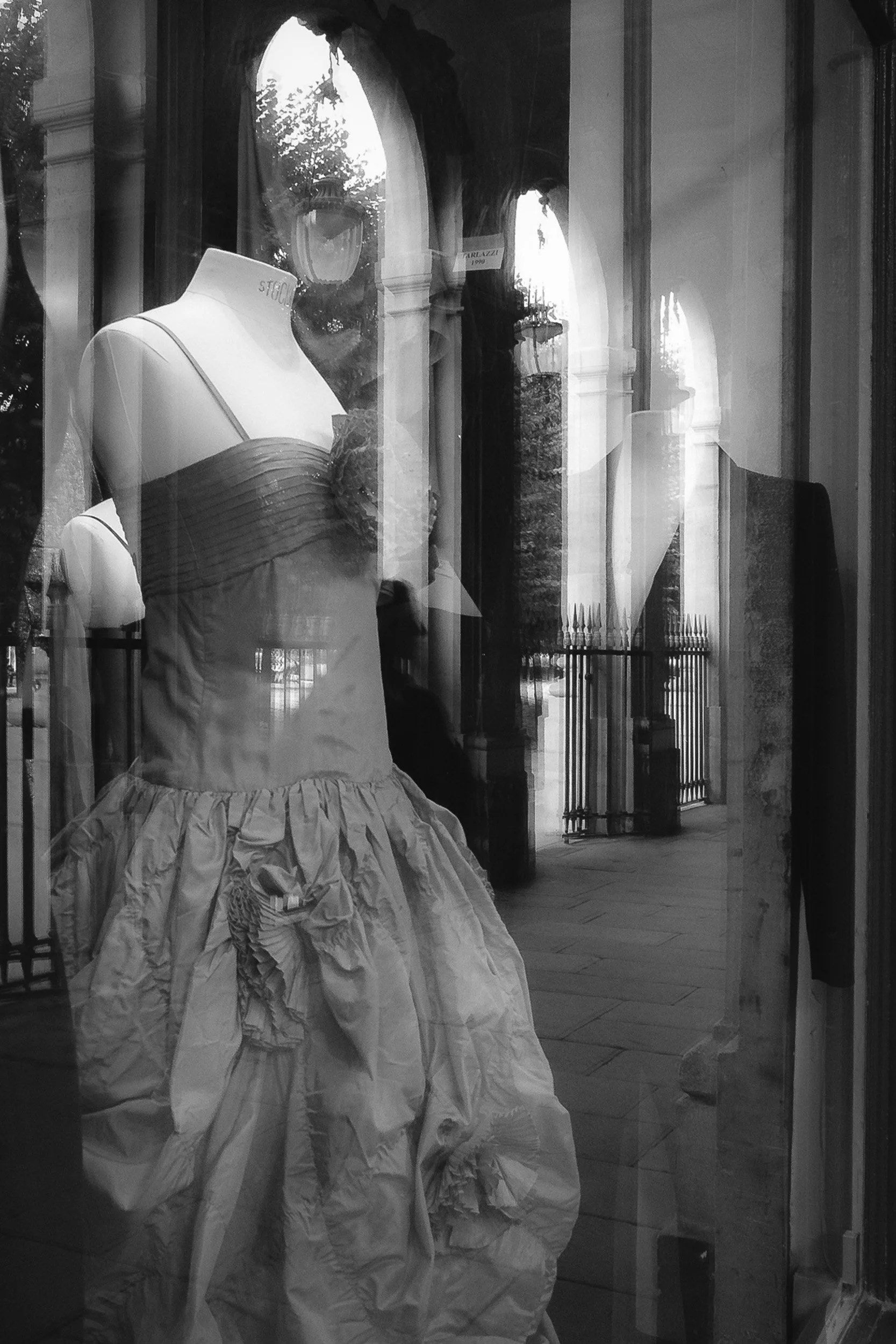 A display window shows a mannequin dressed in an elaborate, voluminous gown with ruffled layers and a strapless bodice, in front of arched windows and ornate building facades outside.
