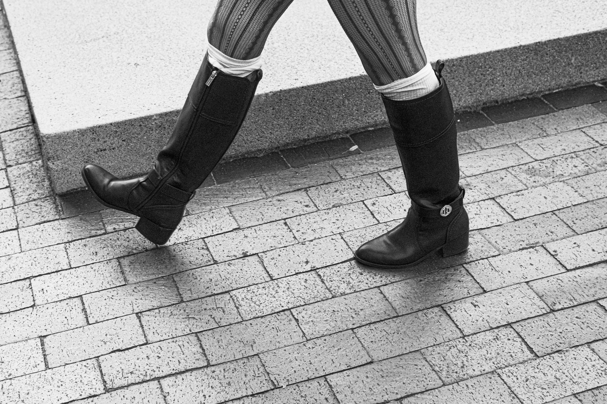 Close-up of a person's legs wearing patterned stockings, white socks, and black knee-high boots with a zipper, standing on a sidewalk with a concrete curb.