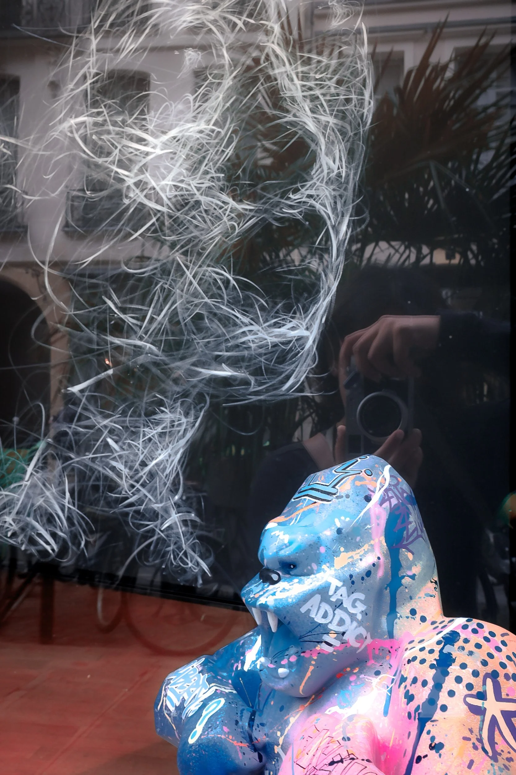 A reflection on a glass surface showing a person wearing a colorful, graffiti-style hoodie with paint splatter designs, a headset, and holding a camera.