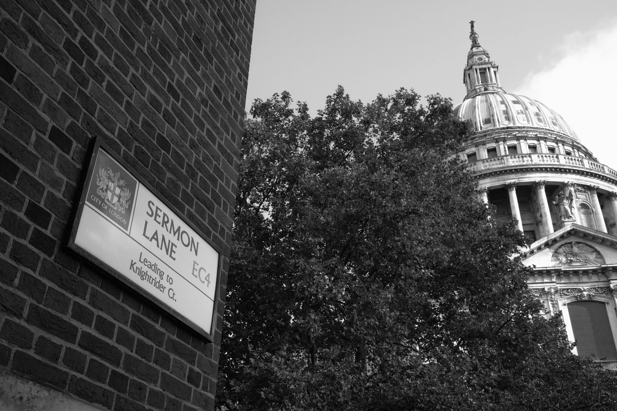 Black and white photo of a city street corner with a brick building on the left and a sign indicating 'Lermon Lane' in London. In the background, part of a historic church or government building with a domed roof and a tall spire is visible, surround