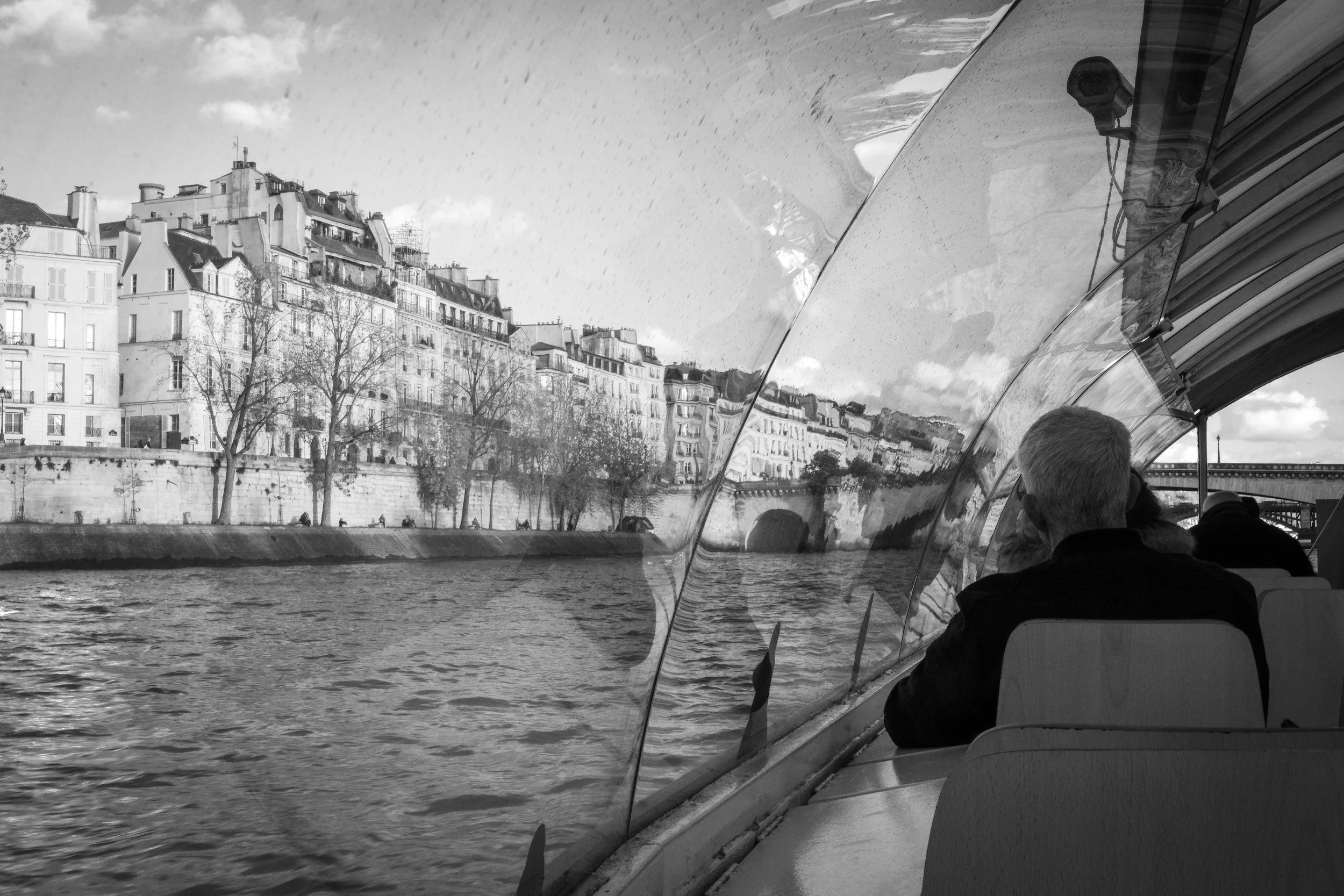 A black and white photo of a riverside cityscape viewed from inside a boat, with people sitting on seats and buildings on the opposite bank.