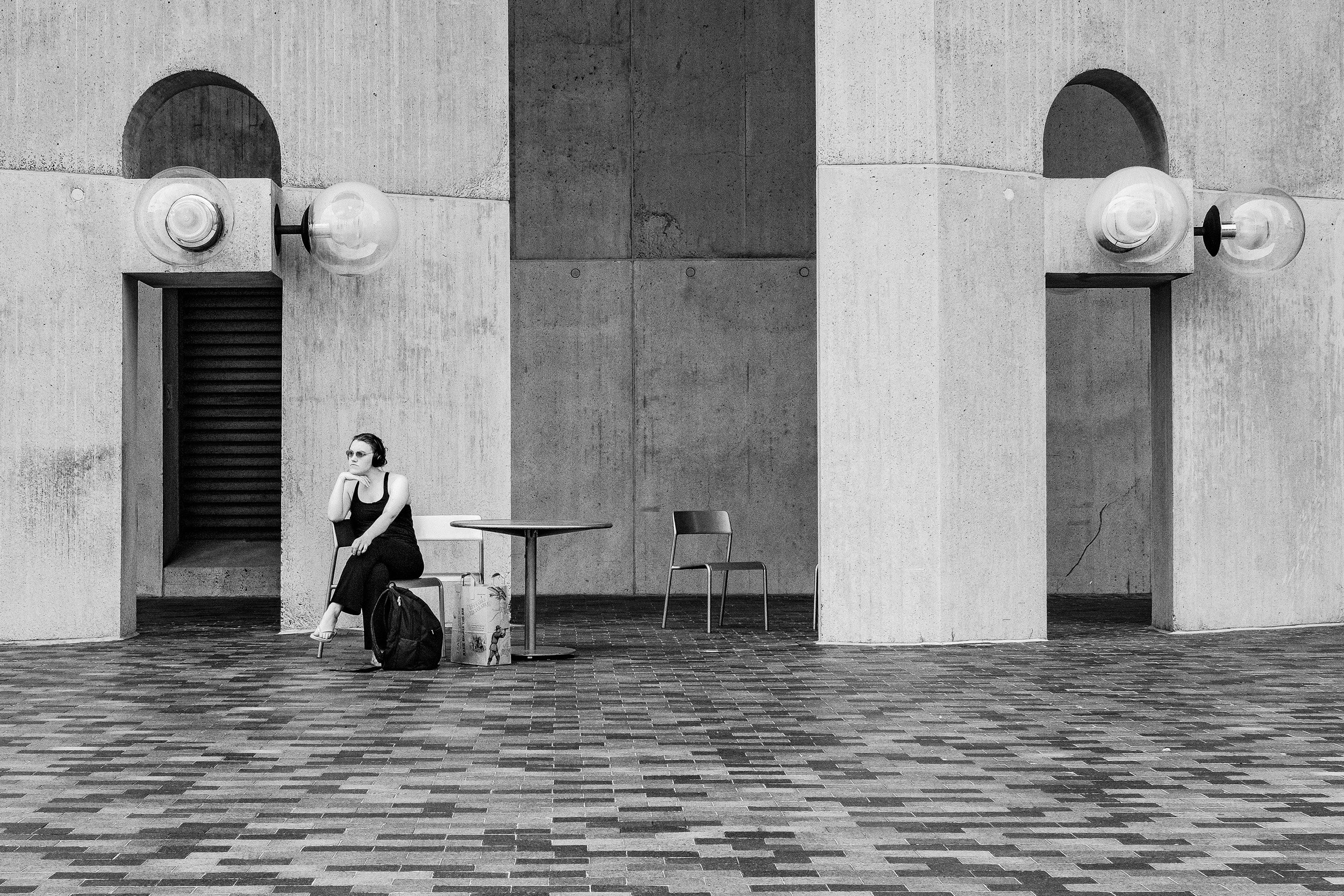 A woman wearing sunglasses and a black dress sitting on a chair with one leg crossed over the other, resting her chin on her hand, next to a table and some luggage, outside against a concrete building with two arched openings and two wall-mounted lig