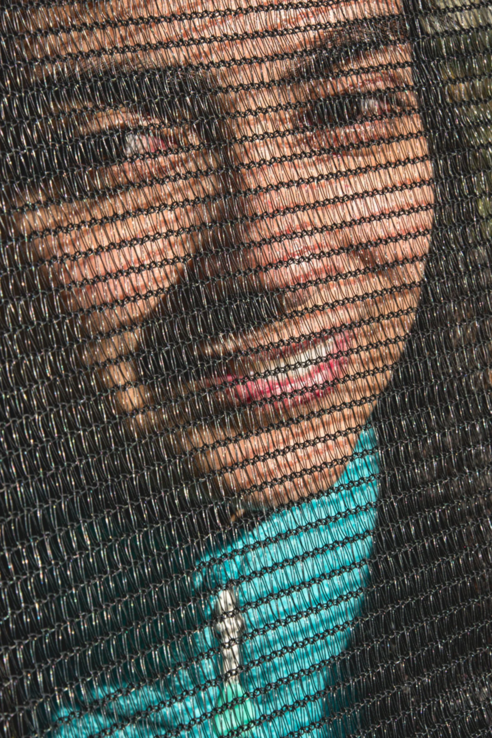 A person's face seen through a black wire mesh screen, with pink lipstick and a turquoise top visible beneath.