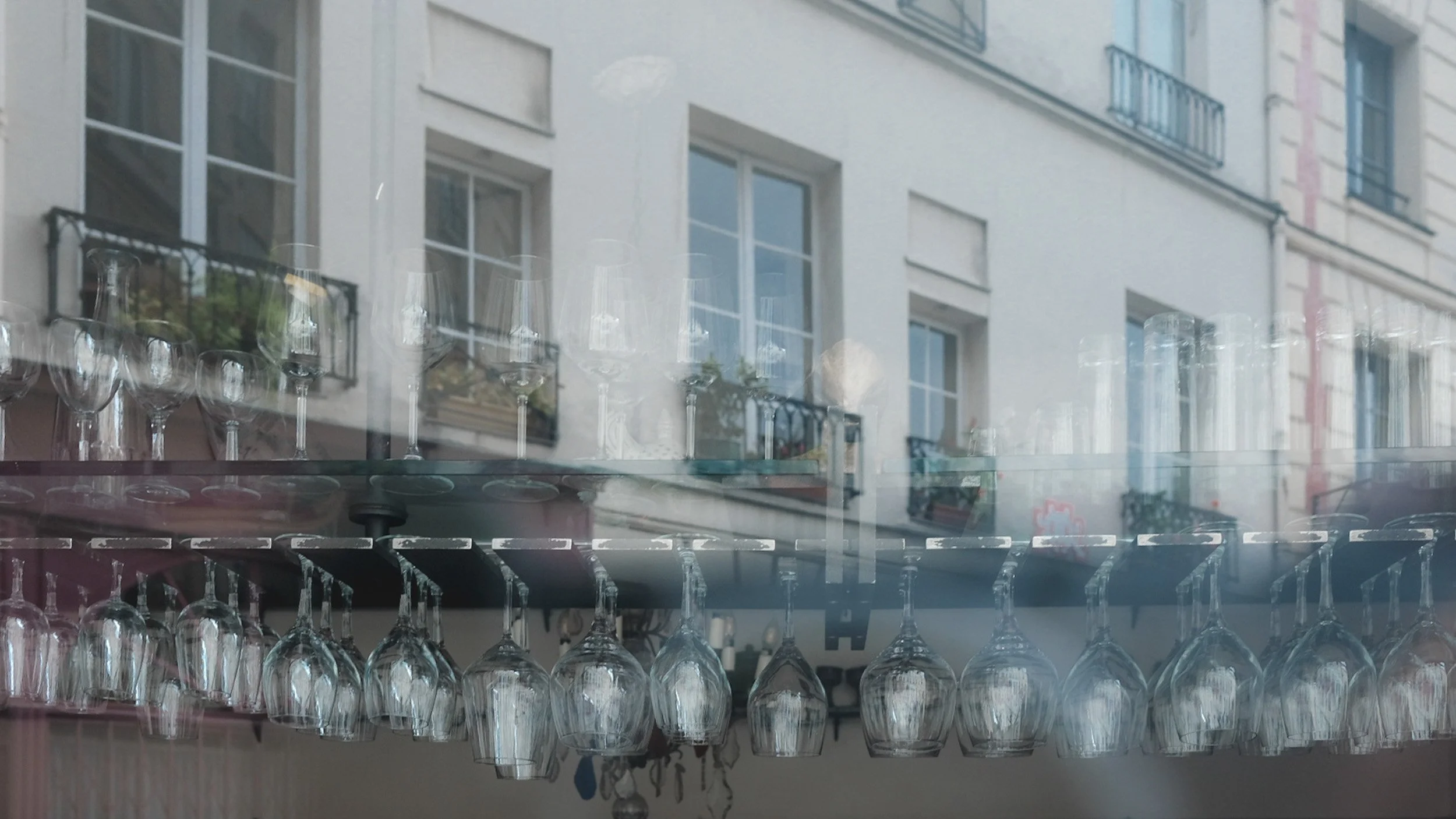 Reflection of a building with multiple windows and balconies seen in a glass display case, with wine glasses hanging upside down.