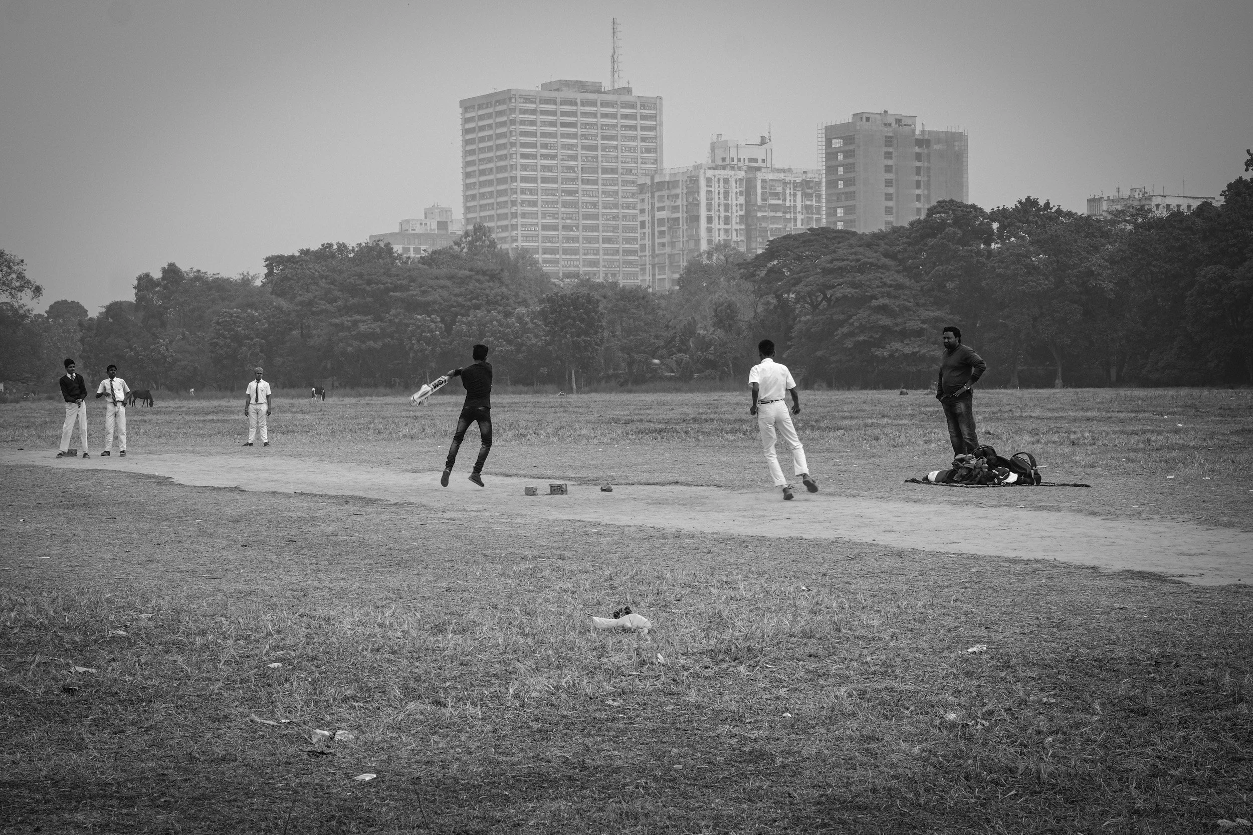 Black and white photo of young men playing cricket on a grassy field with trees and high-rise buildings in the background.