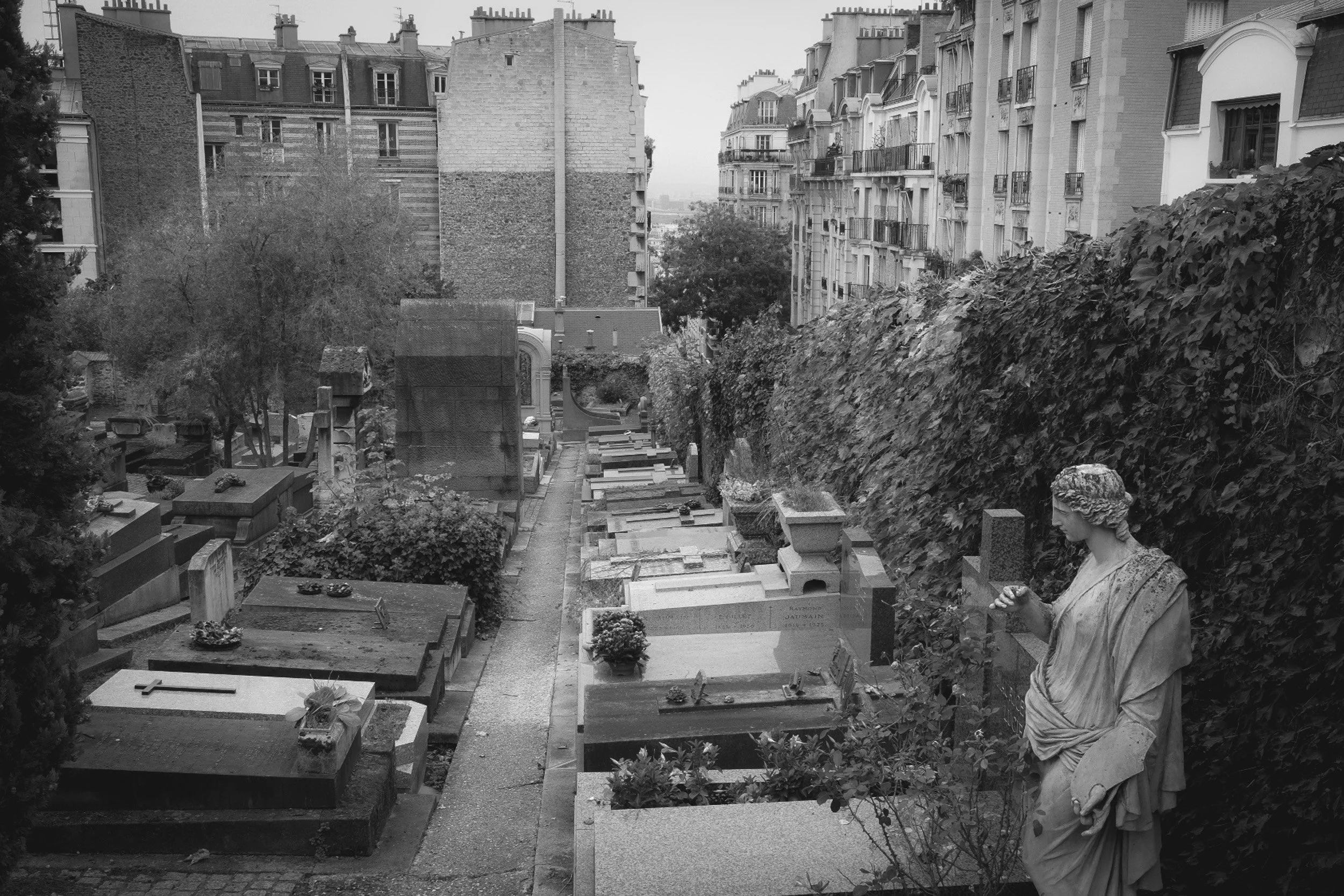 A black and white photo of a cemetery with tombstones and memorials, including a statue of a woman on the right side holding a wreath.