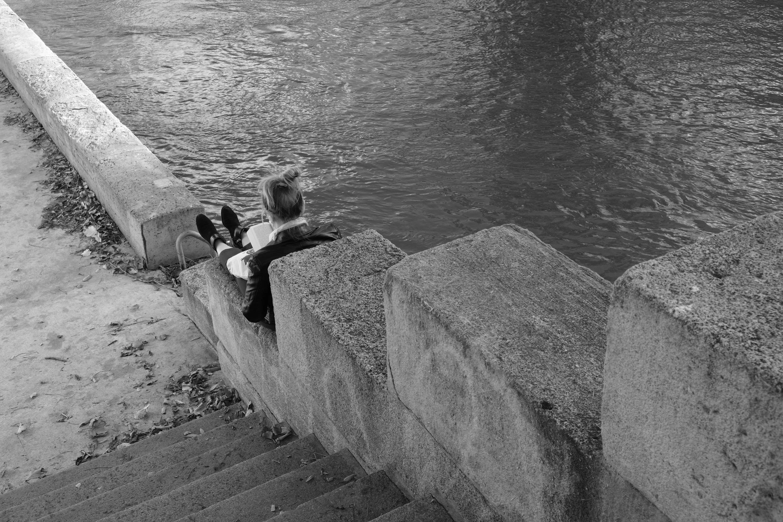 A woman with a ponytail sitting on concrete stairs beside a river, reading a book, with fallen leaves scattered on the ground.