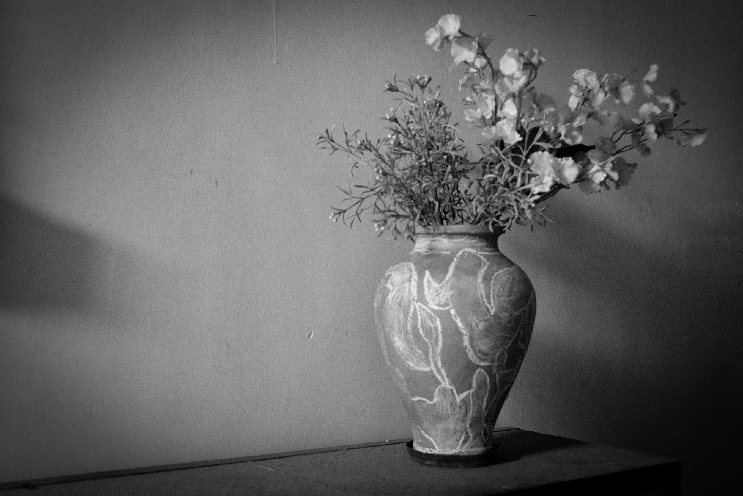 A tall decorative vase with a floral pattern holds a bouquet of small flowers and greenery. The image is in black and white, and the vase is placed on a flat surface against a plain wall.