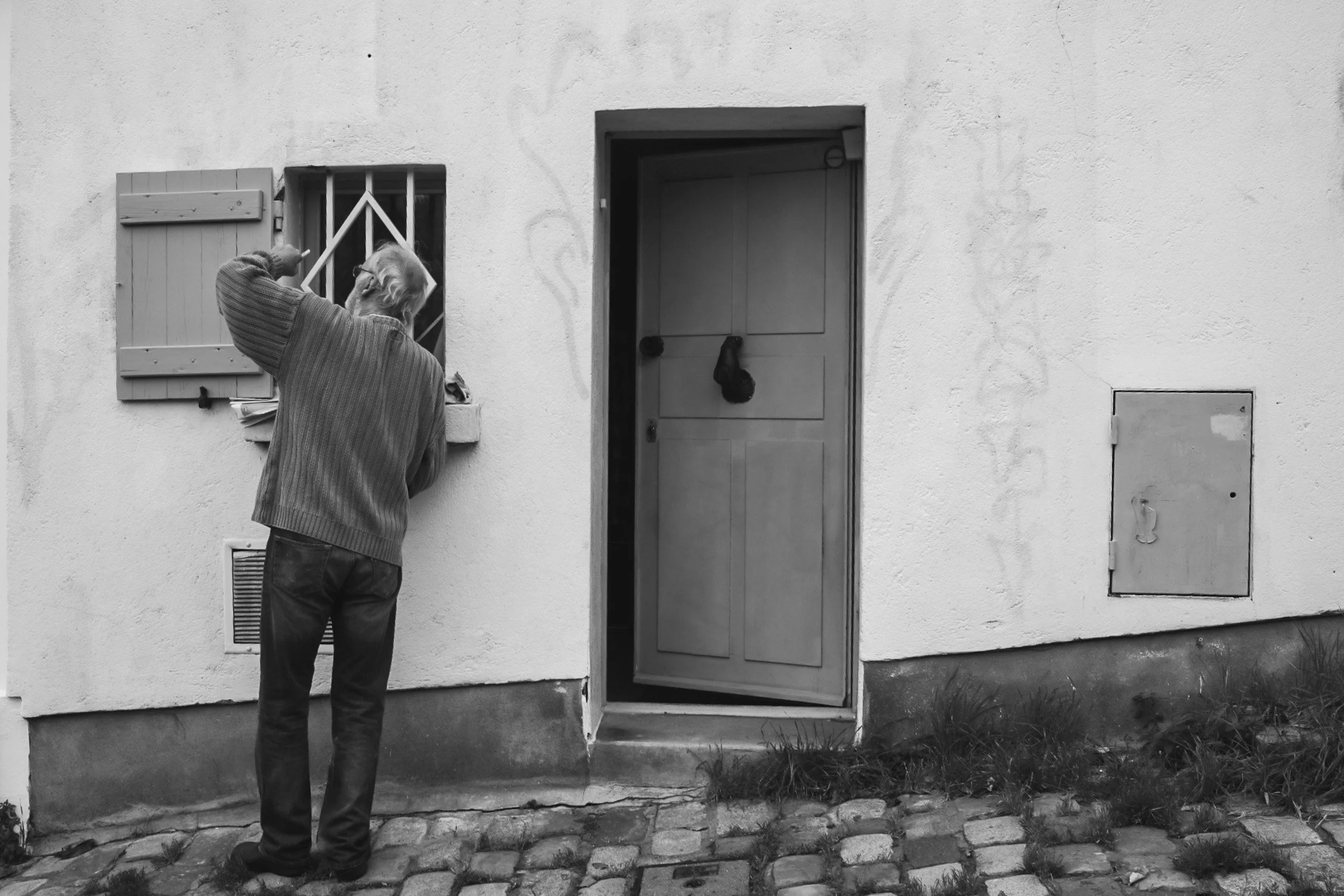 An elderly man with glasses and a striped jacket knocking on a door of a white house.
