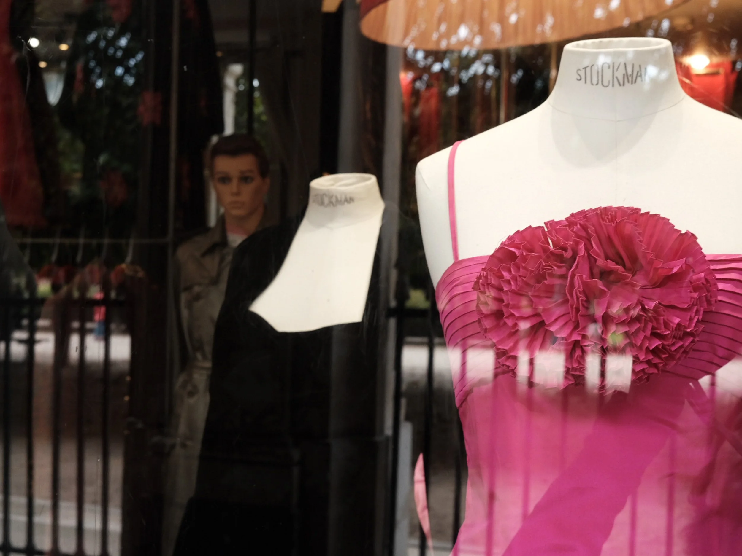 Display window with mannequin dressed in a pink floral dress, behind glass with reflections of the street and a person taking the photo.
