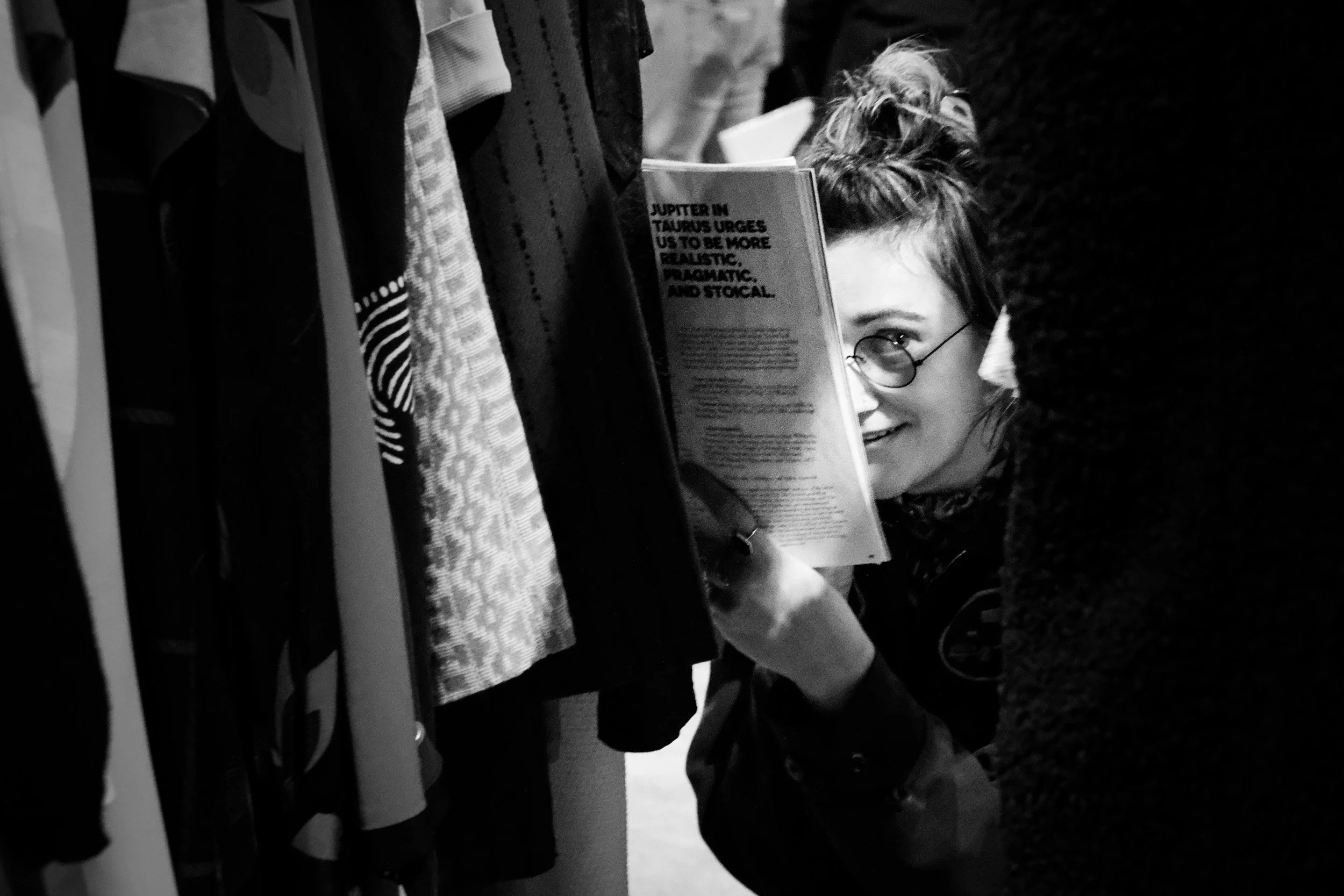 A person with glasses and a ponytail is peeking out from behind a rack of clothes while holding a book that has the visible text 'JUPITER IN TAURUS URGES US TO BE MORE REALISTIC, PRACTIC, AND STROCAL' on the cover.