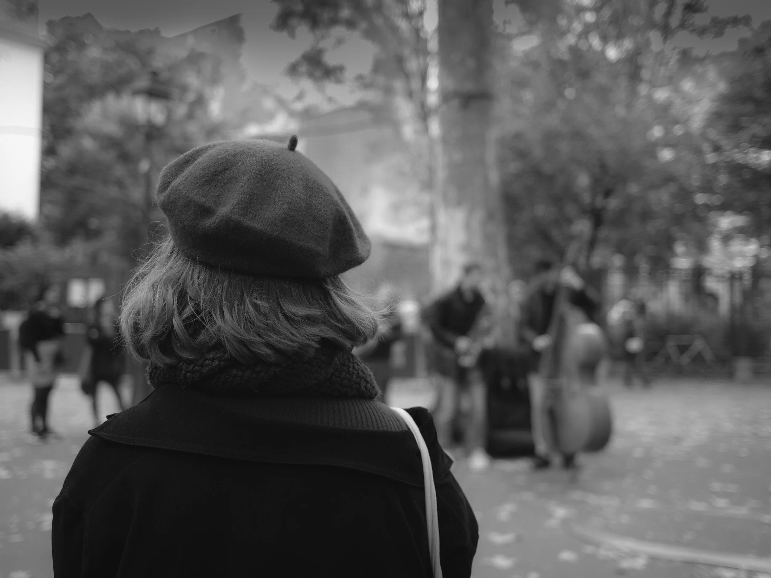 A person wearing a beanie and a jacket looking away in a park, with trees and several people in the background.