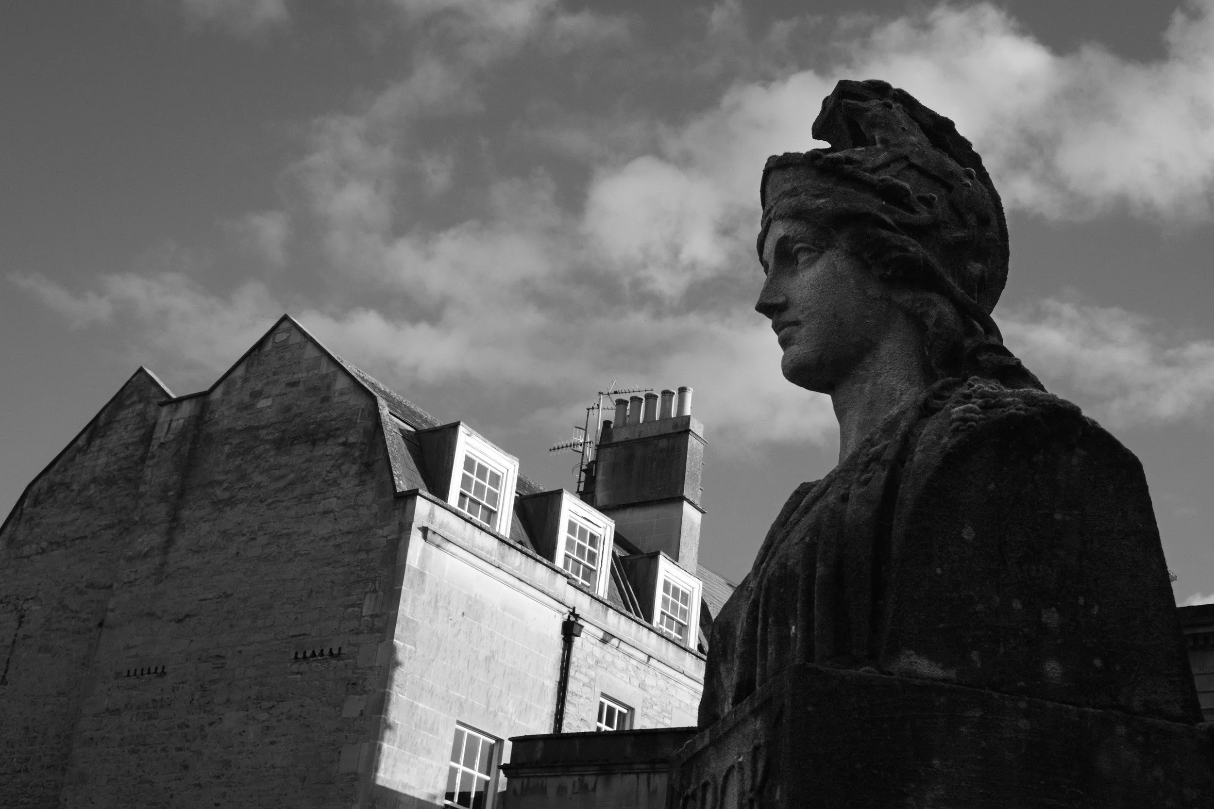 Black and white photo of a stone statue of a person's head with a large headdress, positioned in front of classic European building with multiple chimneys and windows, under cloudy sky.