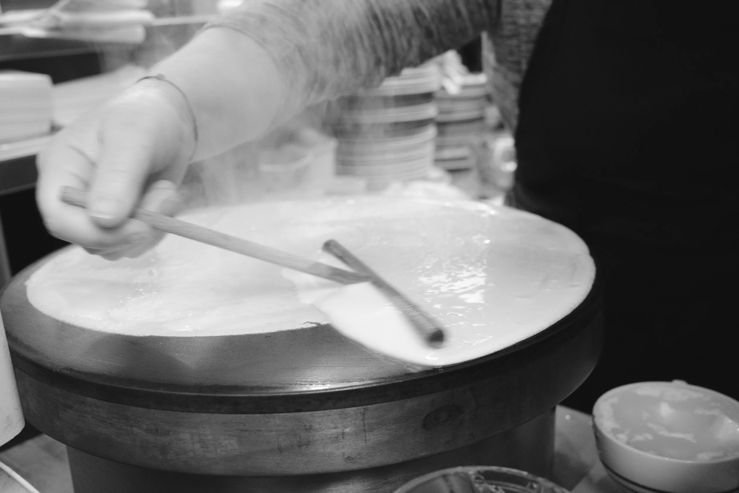 Black and white photo of a person spreading cheese on a pizza in a kitchen or pizzeria setting.