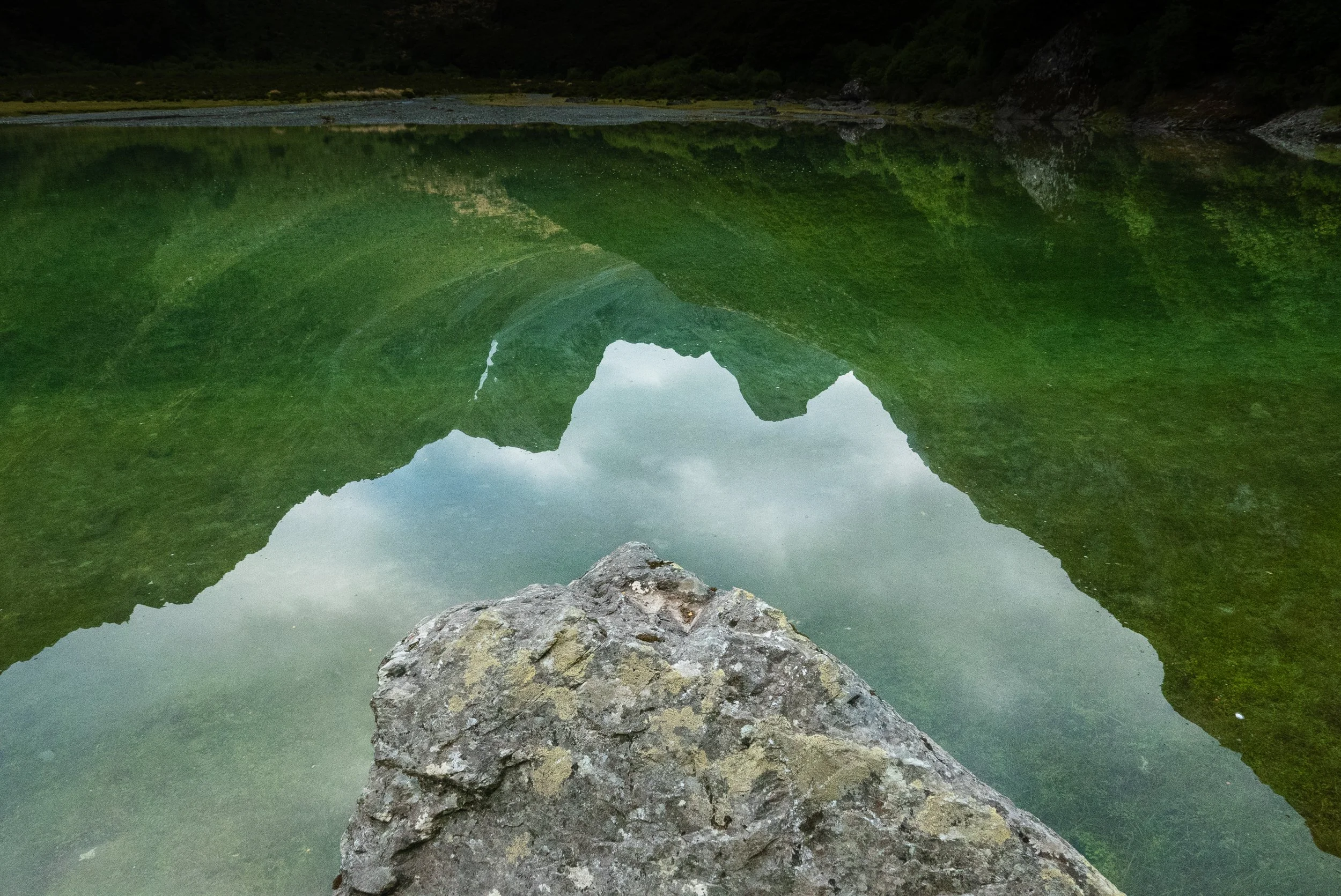 Reflection of a green mountainous landscape in a calm lake with a rocky foreground.