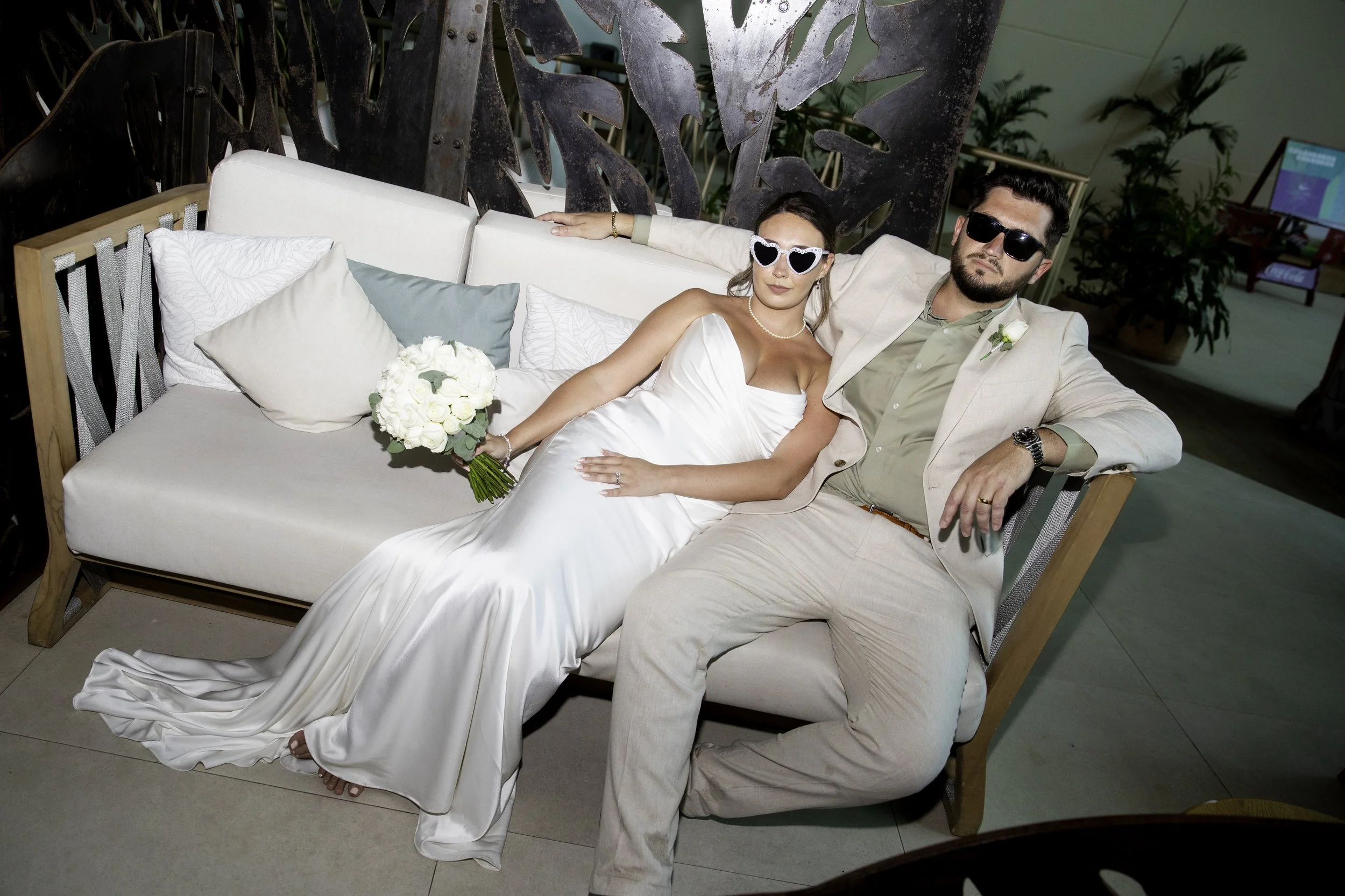 A bride and groom sitting on a white couch in a public indoor space. The bride is holding a bouquet of white roses, wearing a white strapless wedding gown and sunglasses. The groom is dressed in a tan suit with a light green shirt, sunglasses, and a white boutonniere. They are relaxed, leaning back on the couch.