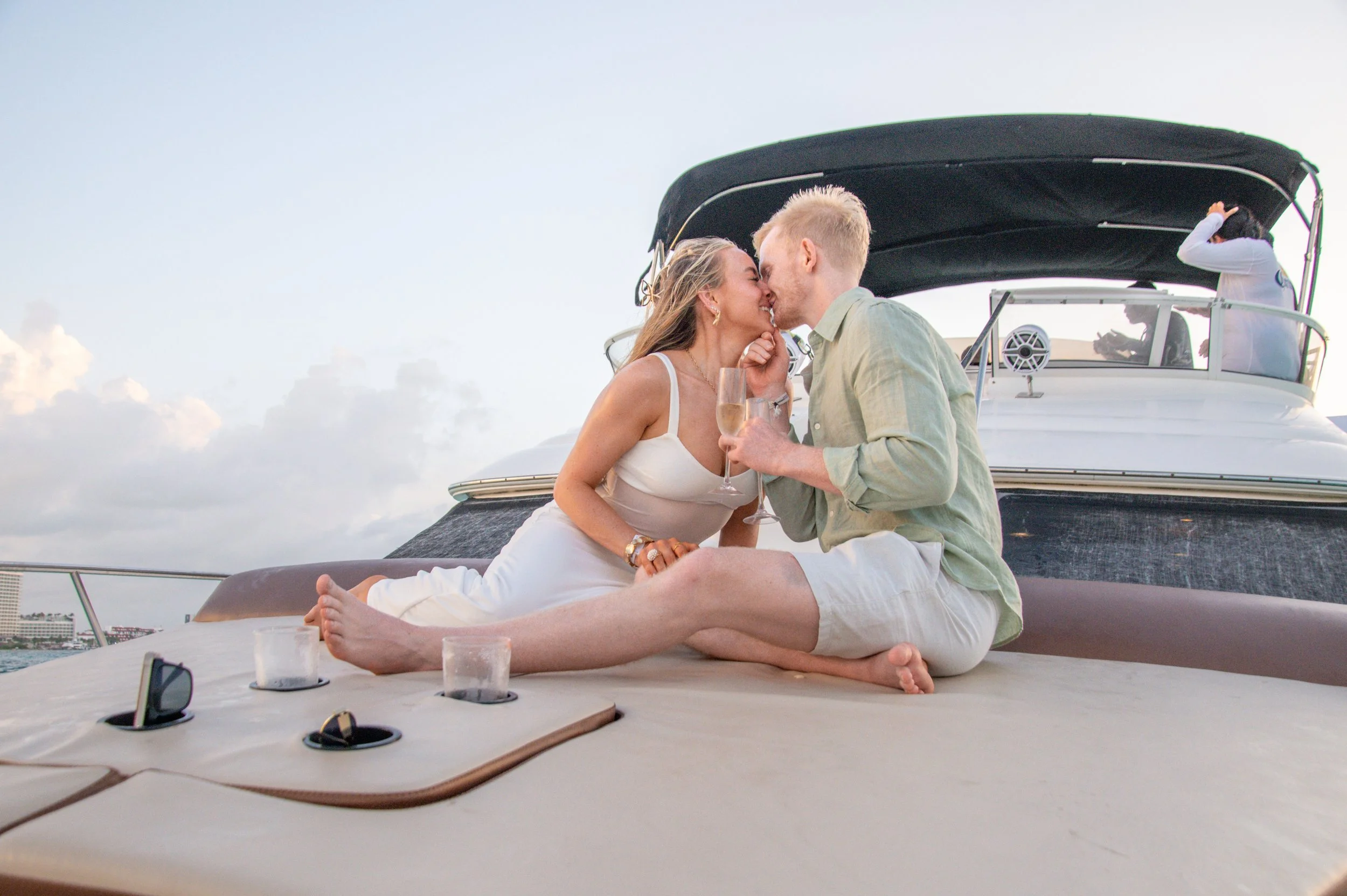 A couple sitting on the deck of a yacht, sharing a tender moment and kissing, with the woman holding a glass of champagne.