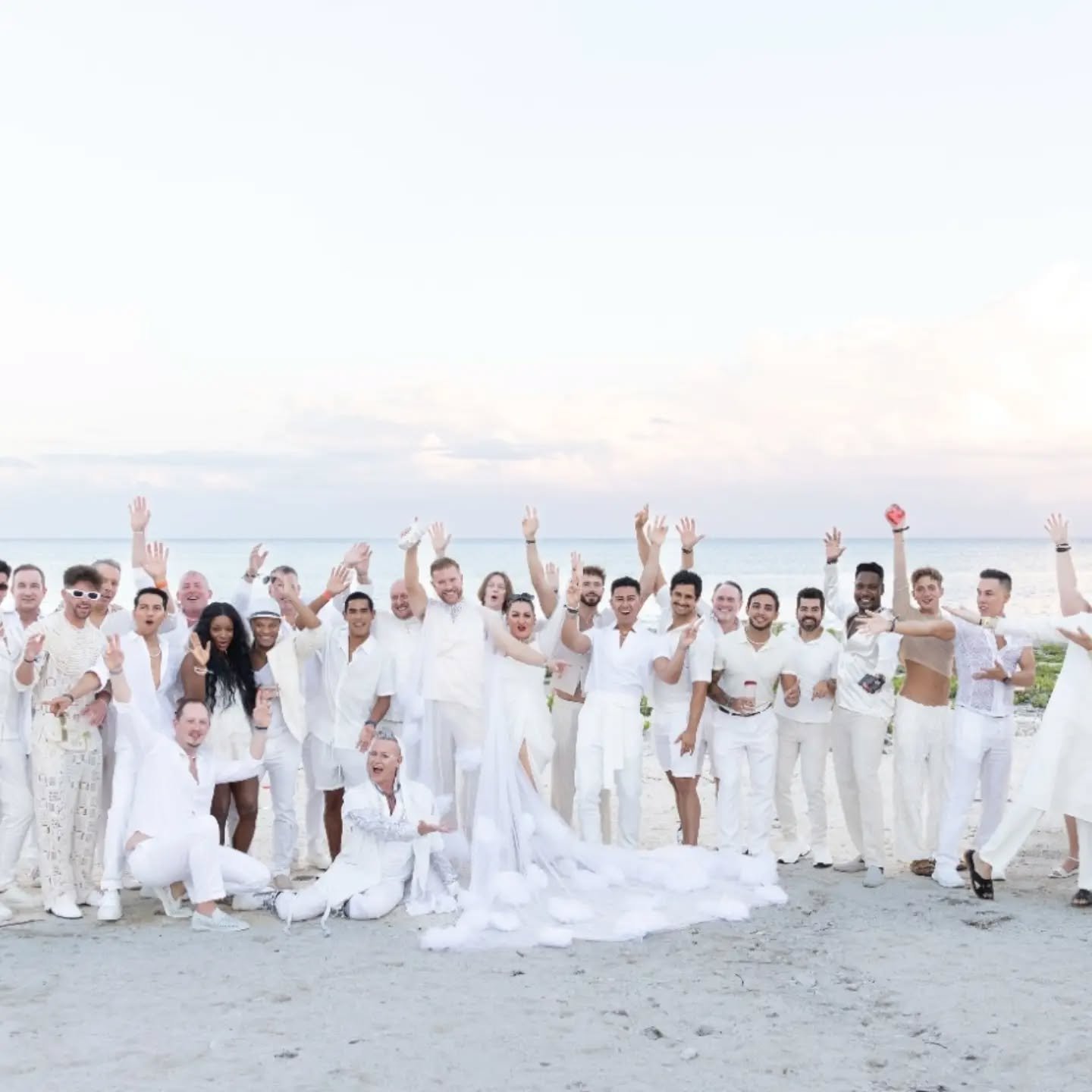 Large group of people dressed in white, celebrating on a beach with the ocean and sky in the background.