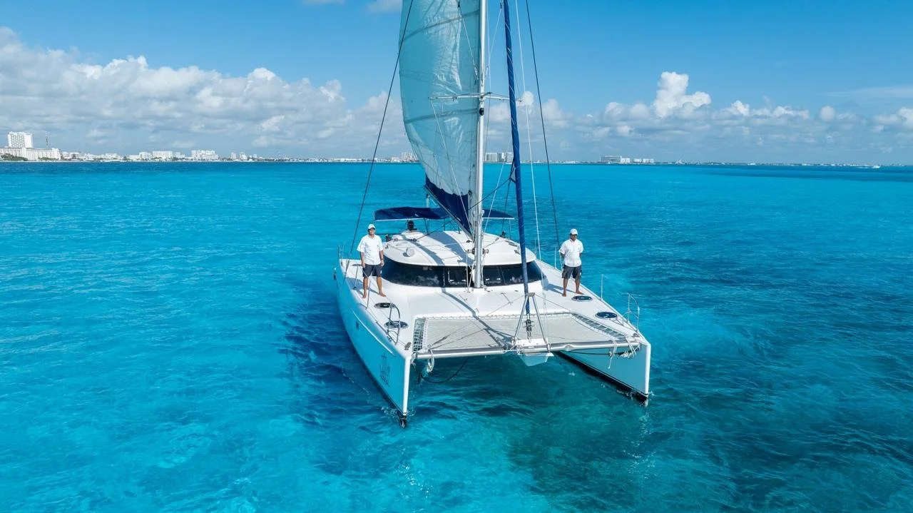 A white sailing catamaran with two men on the deck sailing on clear blue water with a city skyline in the distance under a partly cloudy sky. Catamaran, Cancun, Private vessel, private catamaran