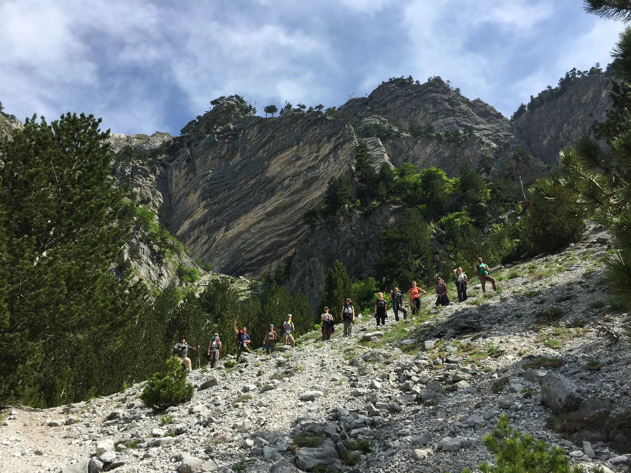 A group of hikers walking up a rocky mountain trail surrounded by green trees, with a large mountain and blue sky in the background.