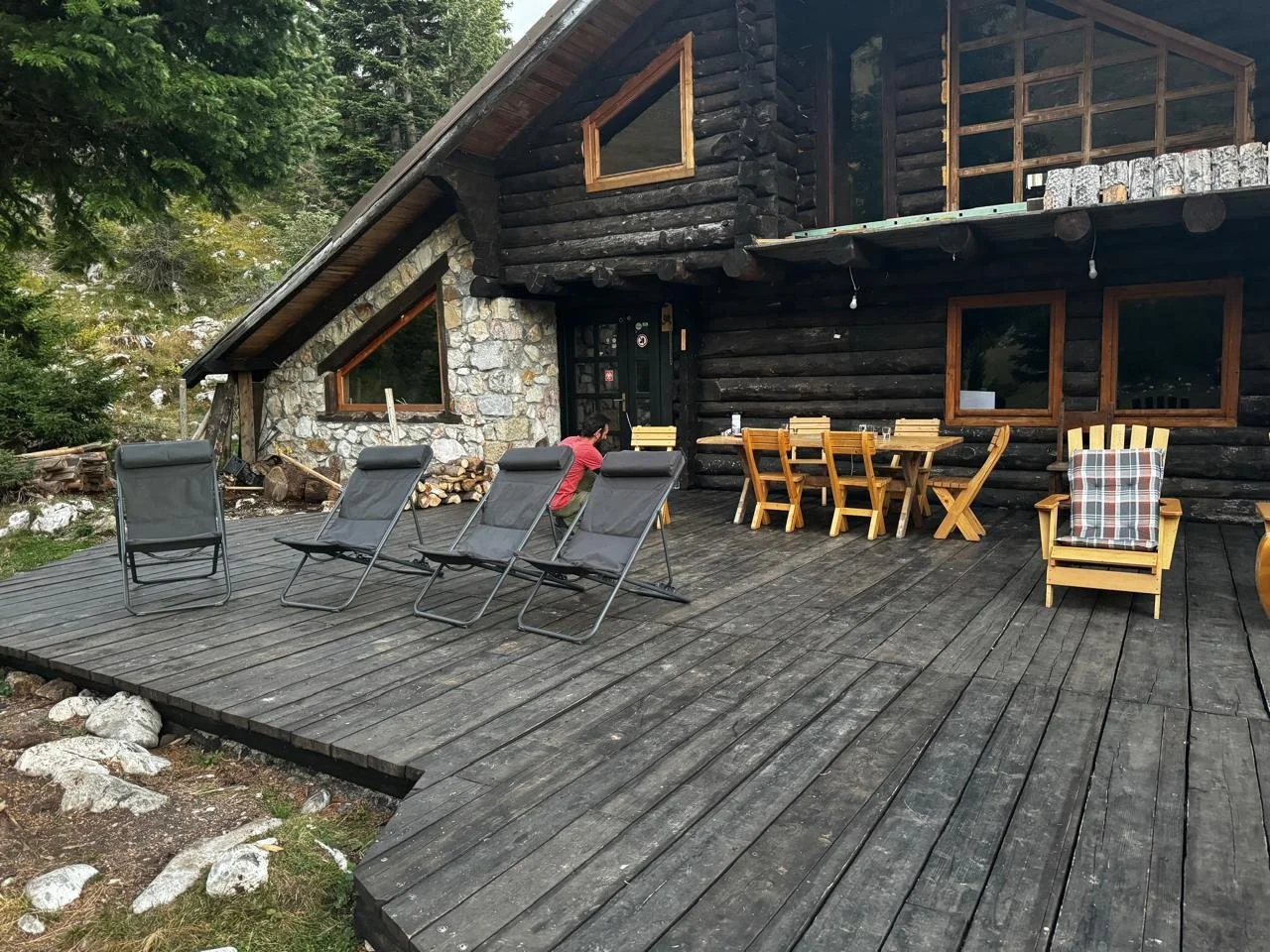 A wooden porch in front of a log cabin with outdoor seating, including four black reclining chairs, a wooden dining table with eight chairs, and a wooden armchair with a plaid cushion. A person in a red shirt is sitting near the house, and trees surr