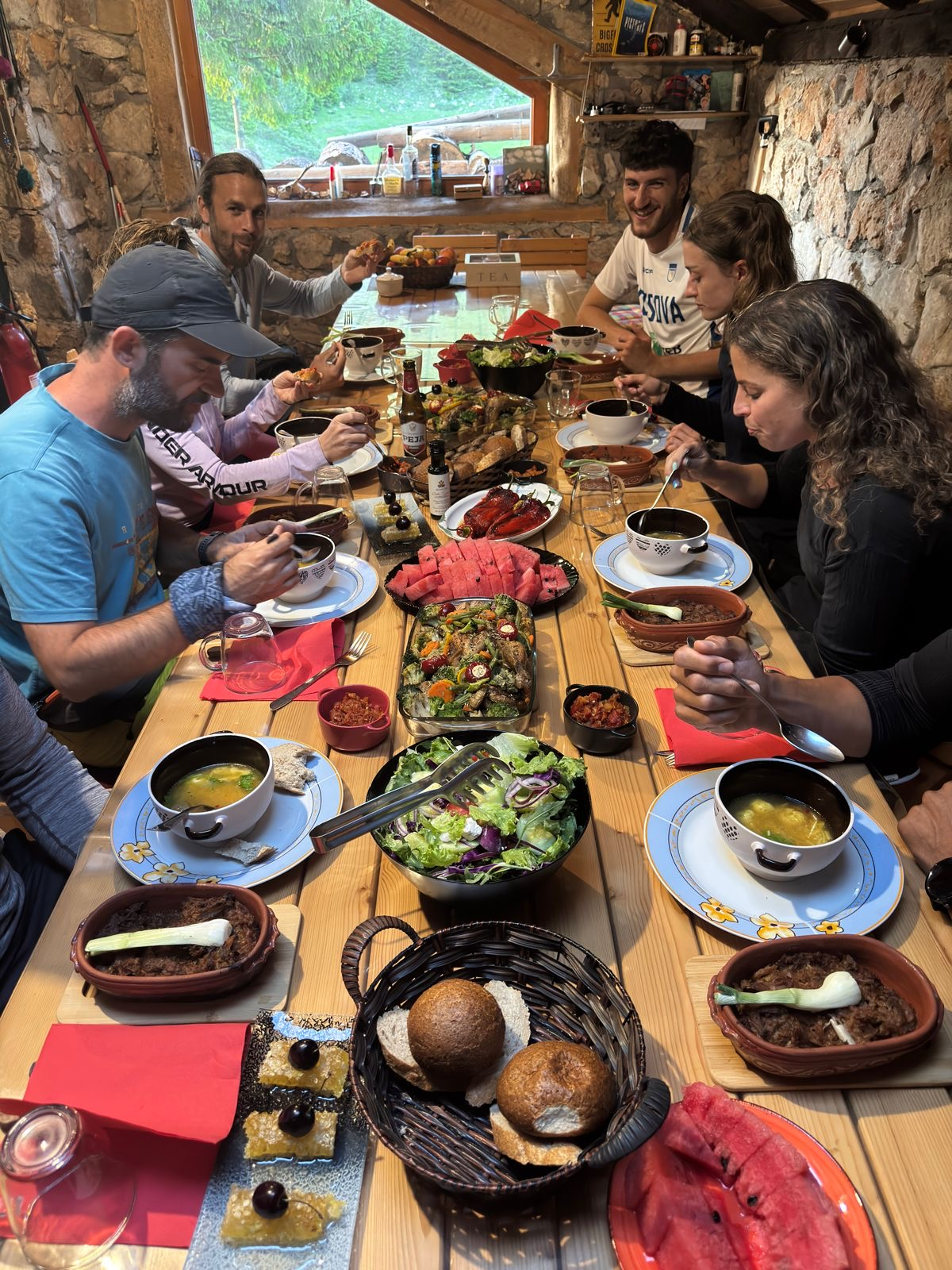 A group of people sitting around a wooden table enjoying a meal in a rustic room with a stone wall and large window, with various dishes including salad, watermelon, bread, and soup.