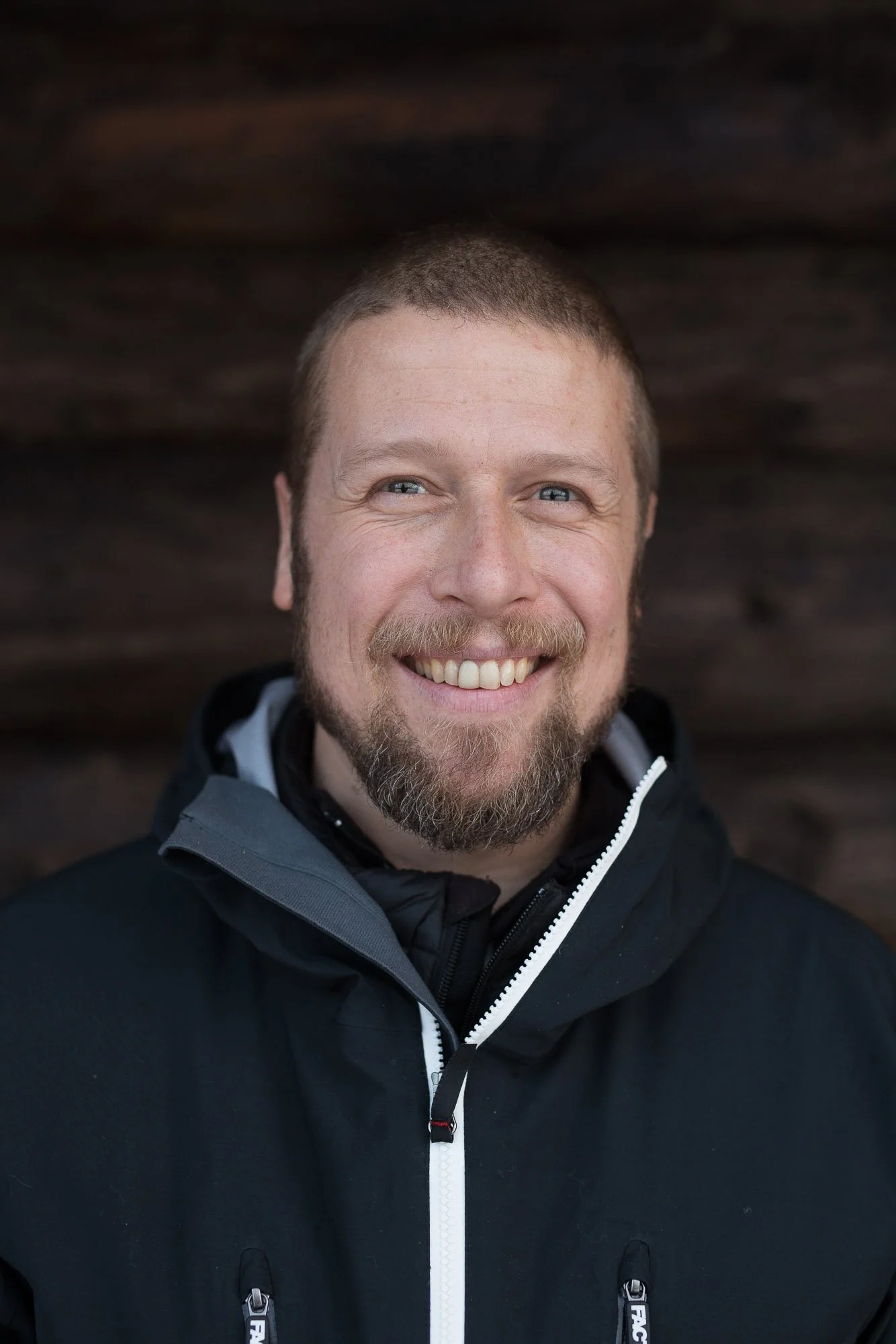 Close-up of a smiling, bearded man with blue eyes and short hair wearing a black jacket with a high collar against a dark wooden background.