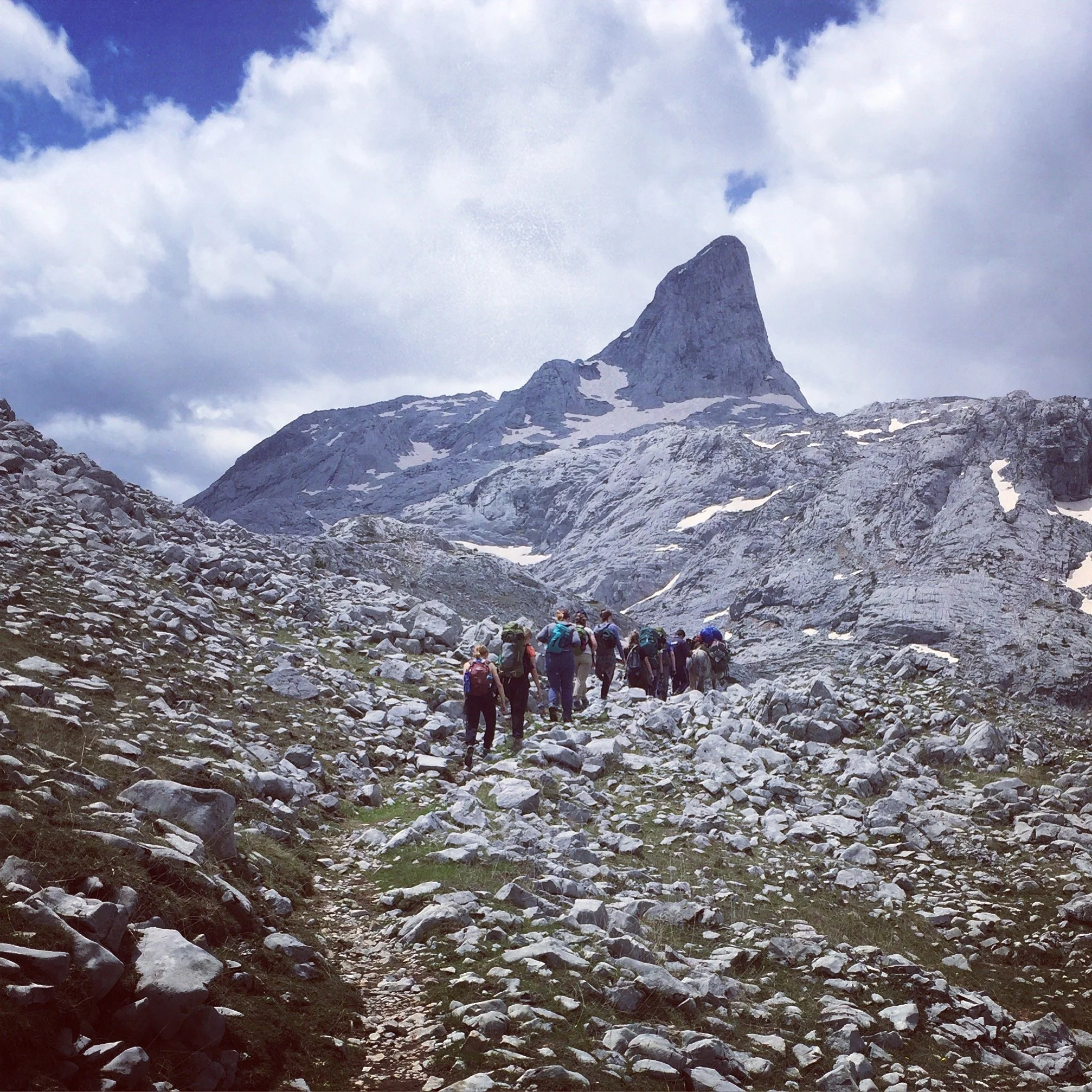 A mountain landscape with a rugged, rocky terrain, snow patches, a mountain peak in the background, and a group of hikers in colorful outdoor clothing walking along a trail.
