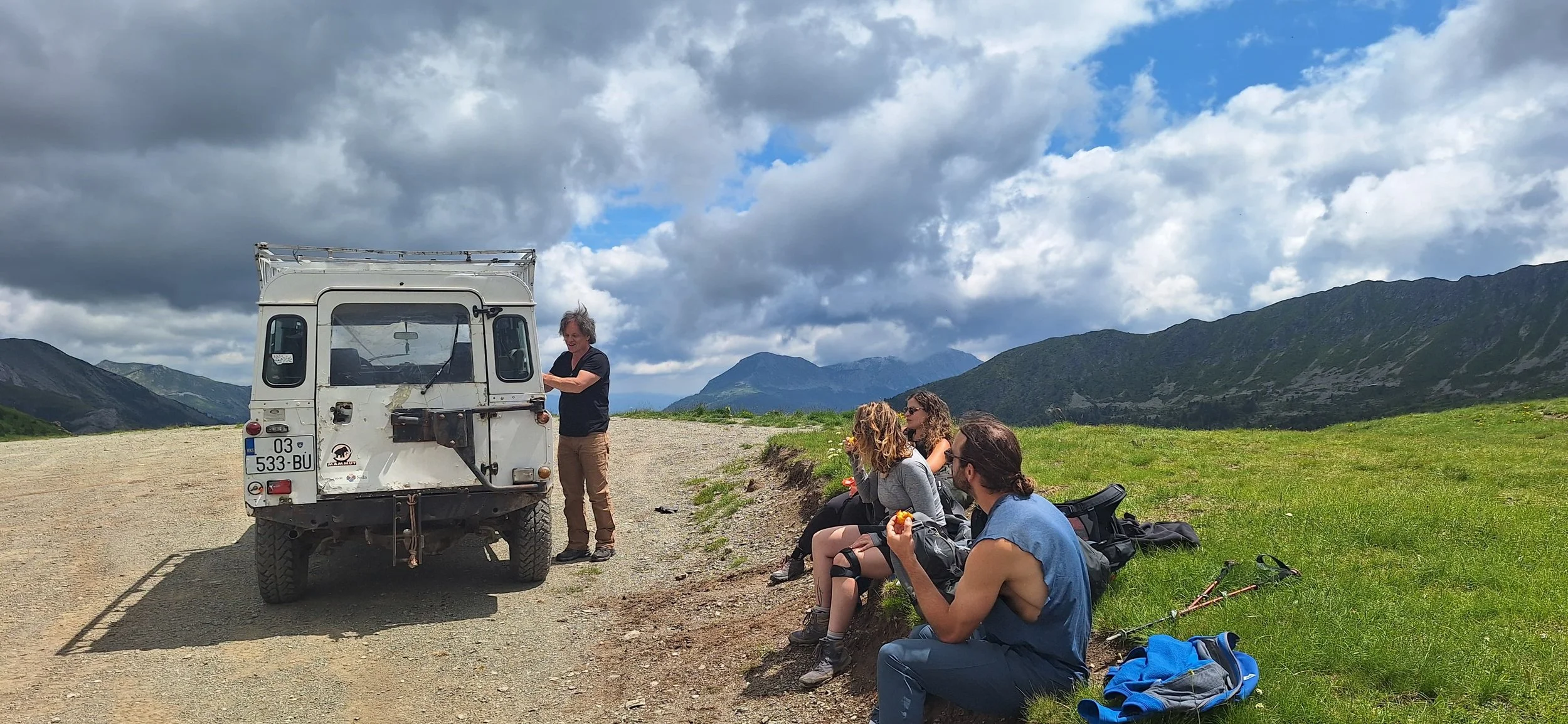 Group of people taking a break with a vintage vehicle on a dirt road in a mountainous landscape, with a cloudy sky.