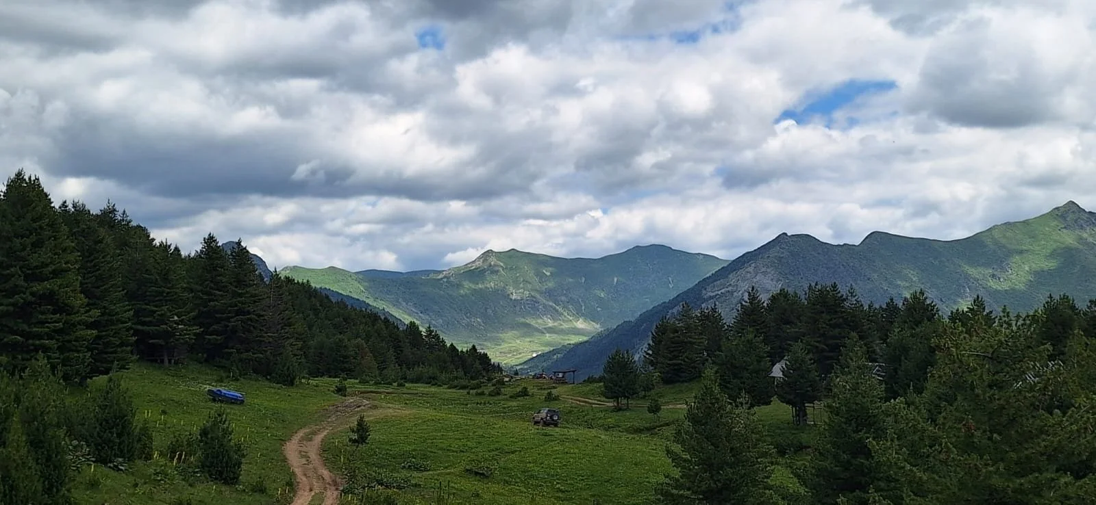 A scenic mountain landscape with green hills, a dirt trail, and trees in the foreground, with mountain peaks under a partly cloudy sky.
