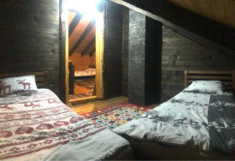 A rustic bedroom with wooden walls, two beds with holiday-themed bedding, and an open door leading to another room with more beds and wooden walls.