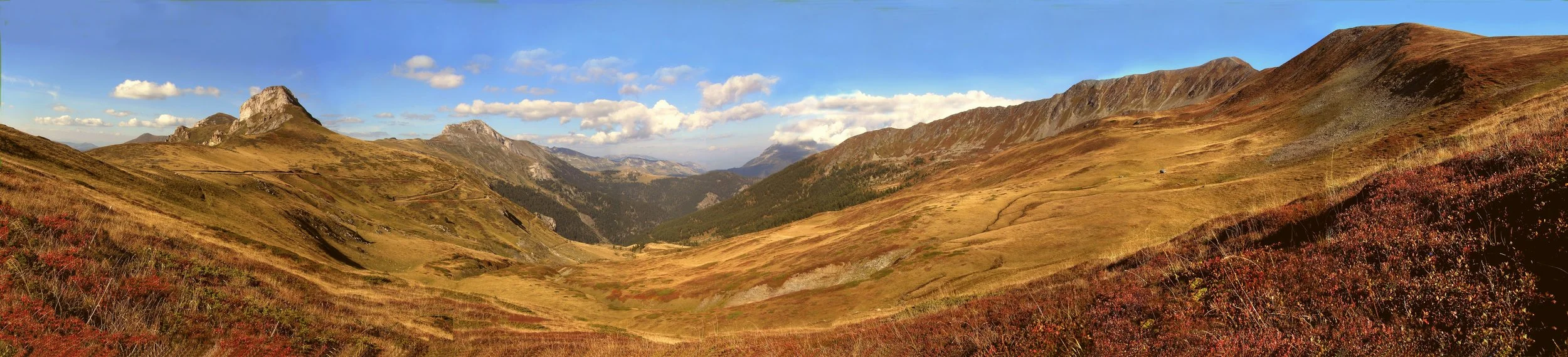 Panoramic view of a mountain landscape with rolling grassy hills and prominent rocky peaks under a partly cloudy sky.