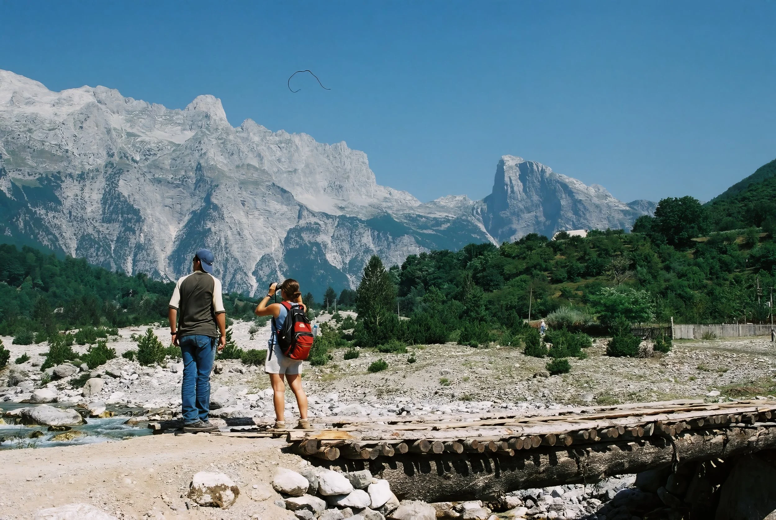 Three hikers, two women and one man, walking in a rocky outdoor area with a mountain range covered in snow in the background. The sky is clear and blue.