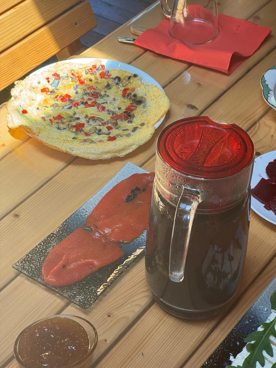 A table set with a slice of bread topped with cheese and chopped vegetables, a glass of iced tea, a small bowl of dipping sauce, and slices of ham on a black rectangular plate.