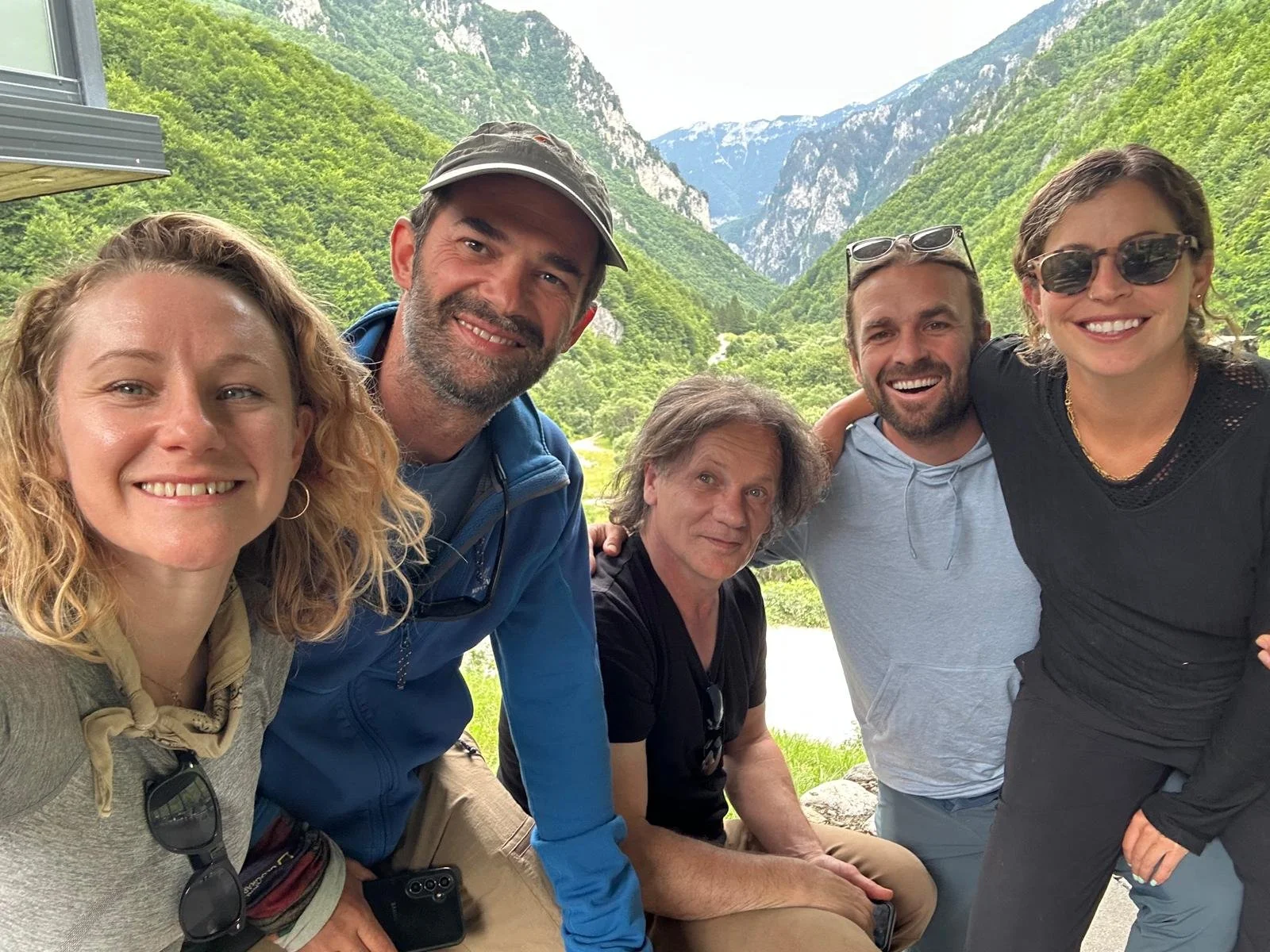 A group of six friends smiling outdoors in a mountainous area with green hills and snow-capped peaks in the background.