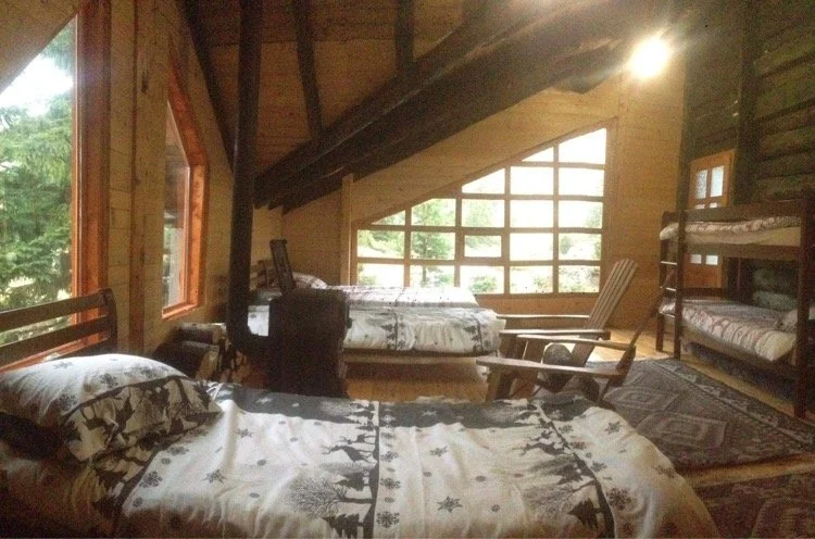 A rustic bedroom with wooden walls and beams, featuring a single bed with holiday-themed bedding, a bunk bed, and a small wooden desk near large wraparound windows letting in natural light.
