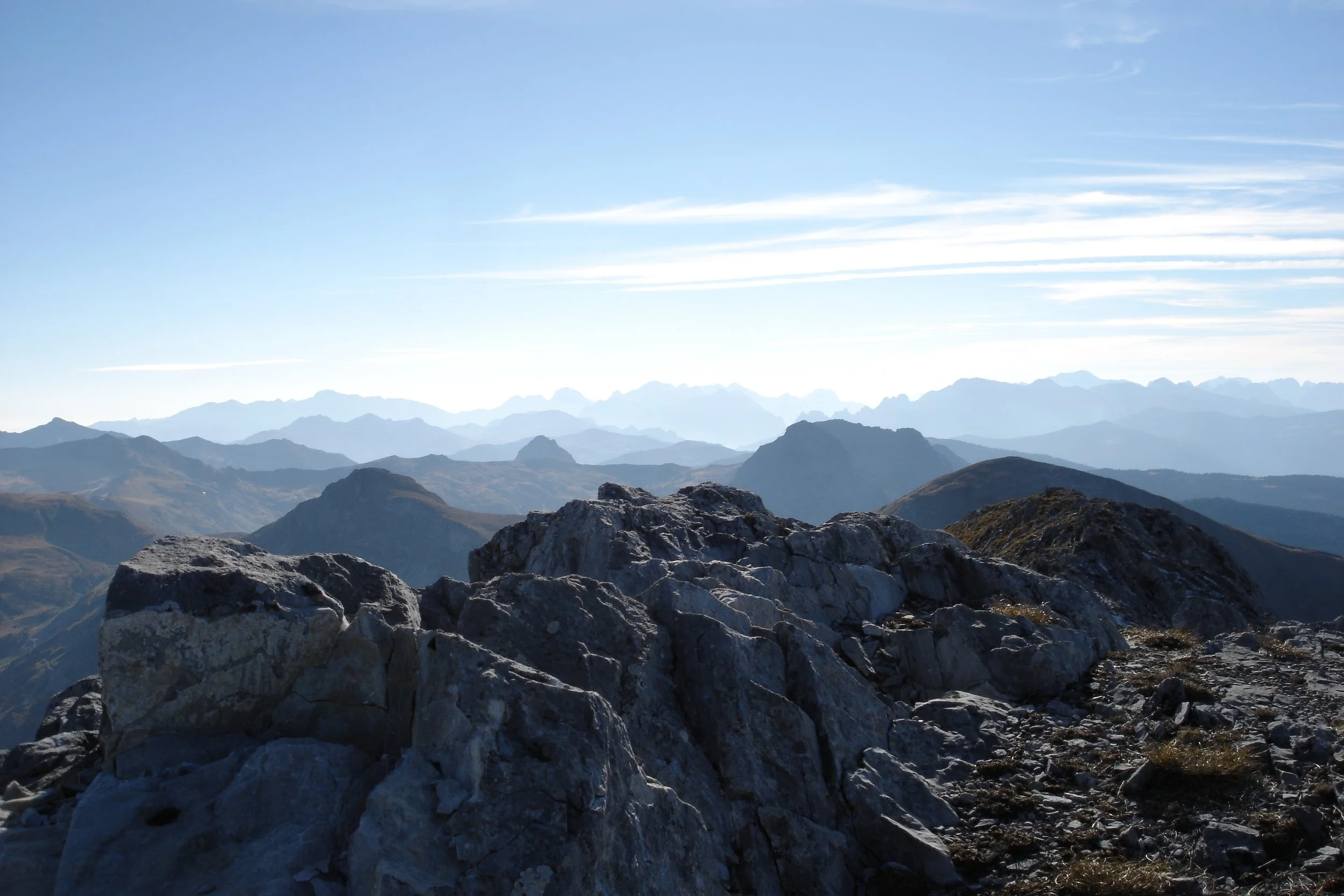A mountain landscape with rocky foreground, multiple mountain ridges in the distance, and a clear blue sky with high wispy clouds.