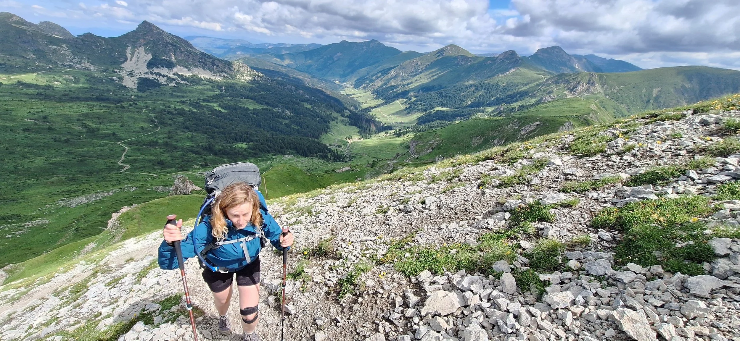 A woman hiking up a rocky mountain trail with a scenic green valley and mountains in the background on a partly cloudy day.