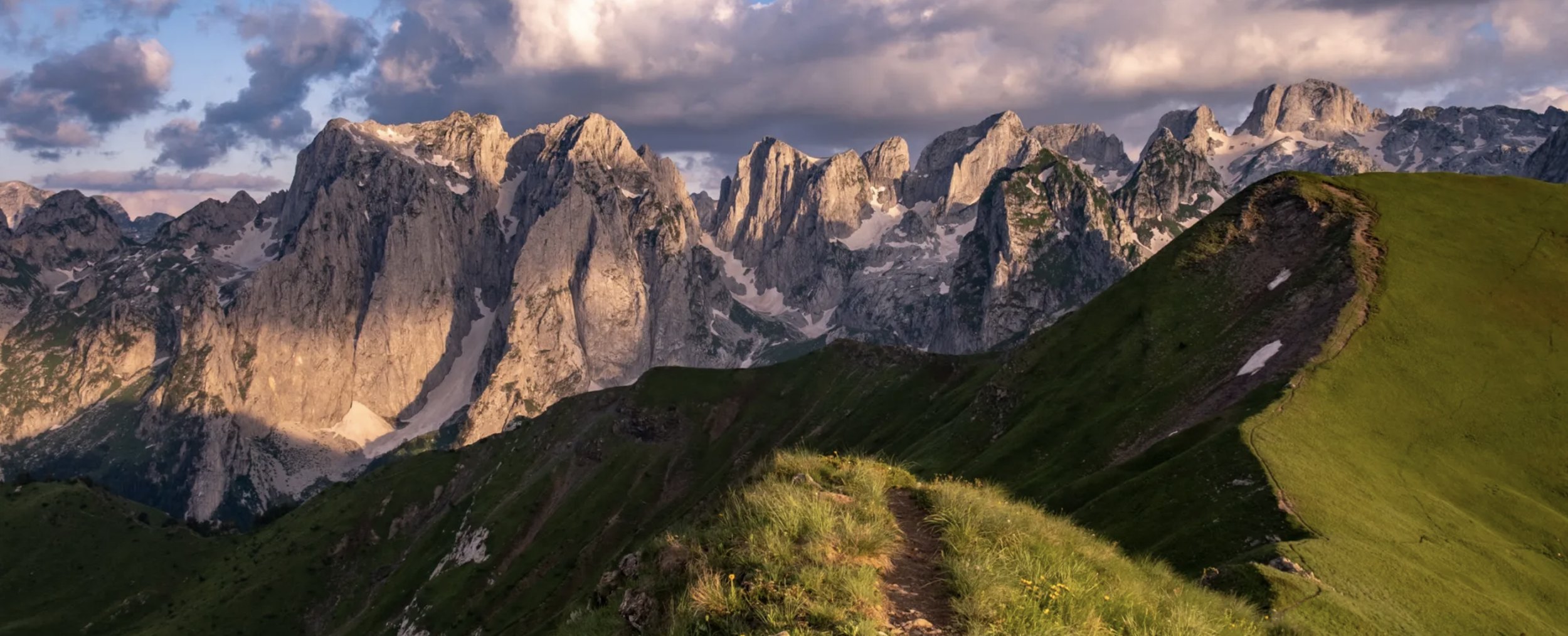 Mountain range with rugged peaks and patches of snow, illuminated by soft sunlight, with green grassy hills and a dirt trail in the foreground under a partly cloudy sky.