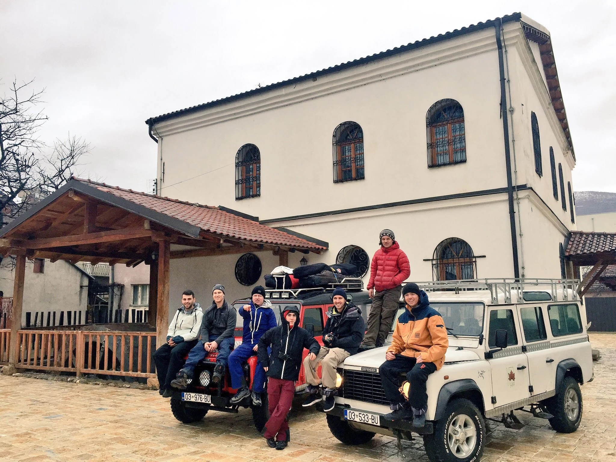 A group of people, mostly young men, posing on the hood of a white off-road vehicle with Spanish license plates, in front of a large beige building with arched windows and black iron bars. Some are standing, others sitting, and one person is standing on the vehicle's hood. The scene appears to be outdoors on a cloudy day with leafless trees in the background.