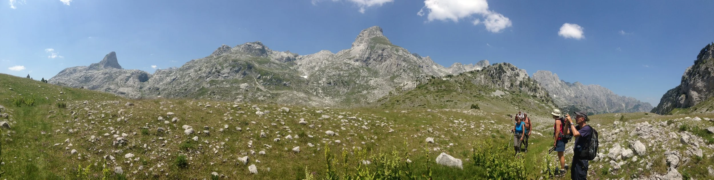 A group of hikers walking through a mountain valley with tall, rocky peaks in the background, under a blue sky with a few clouds.