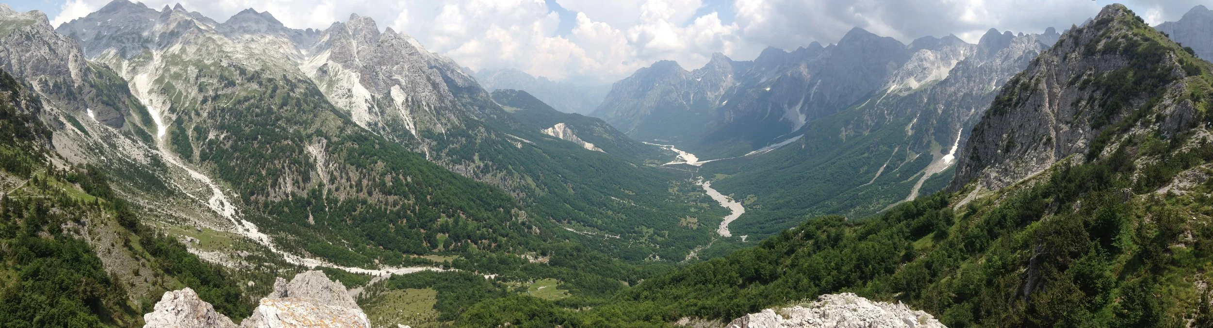 A vast mountain valley with rugged peaks, green forested slopes, and a winding river.