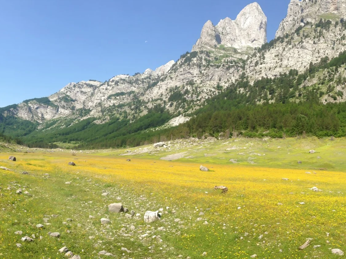 Mountain landscape with rugged peaks, some snow patches, and a green hillside with a trail in the foreground under a partly cloudy sky.